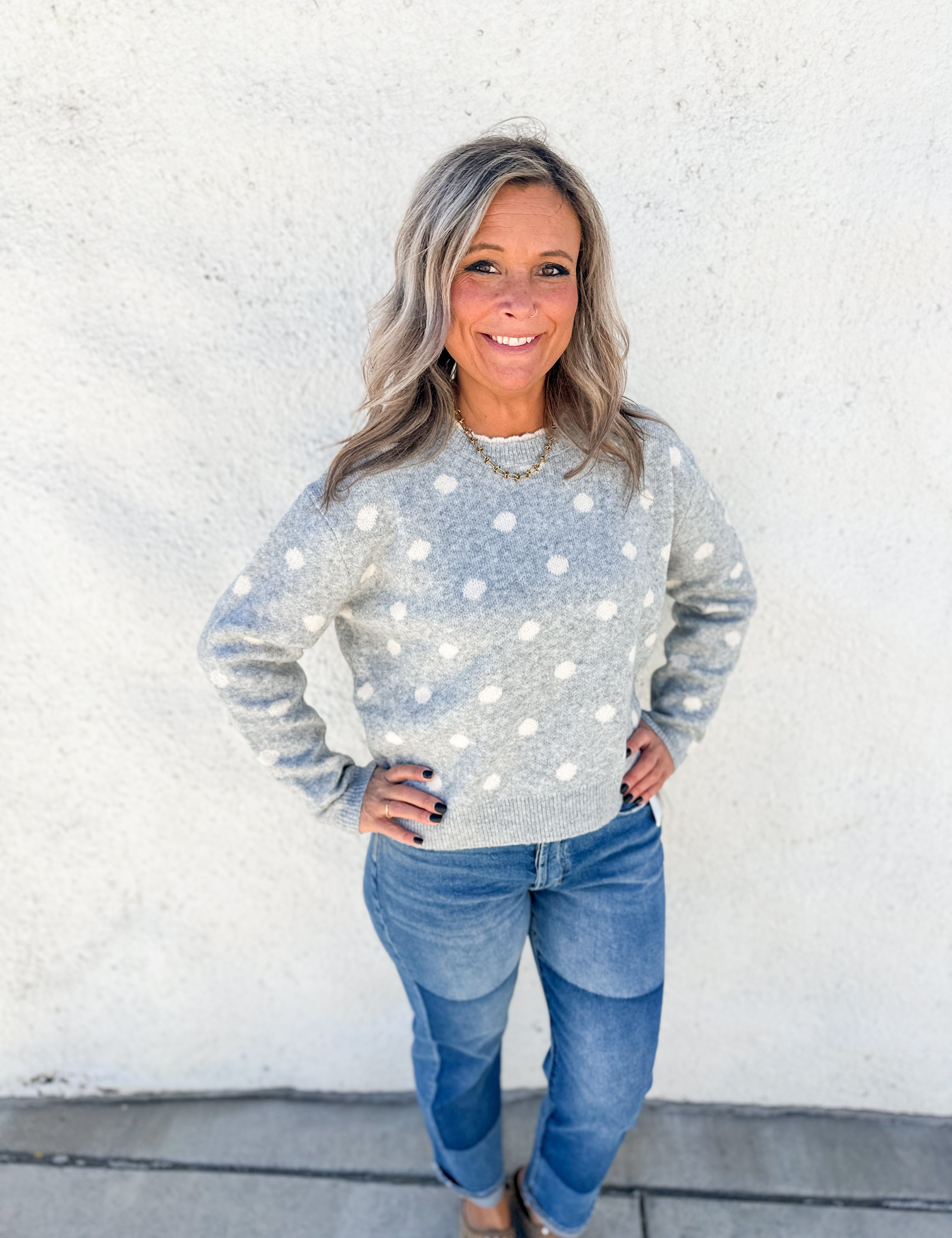 A woman with gray hair stands against a white stucco wall wearing a light gray long-sleeve knit sweater with white polka dots and blue jeans.