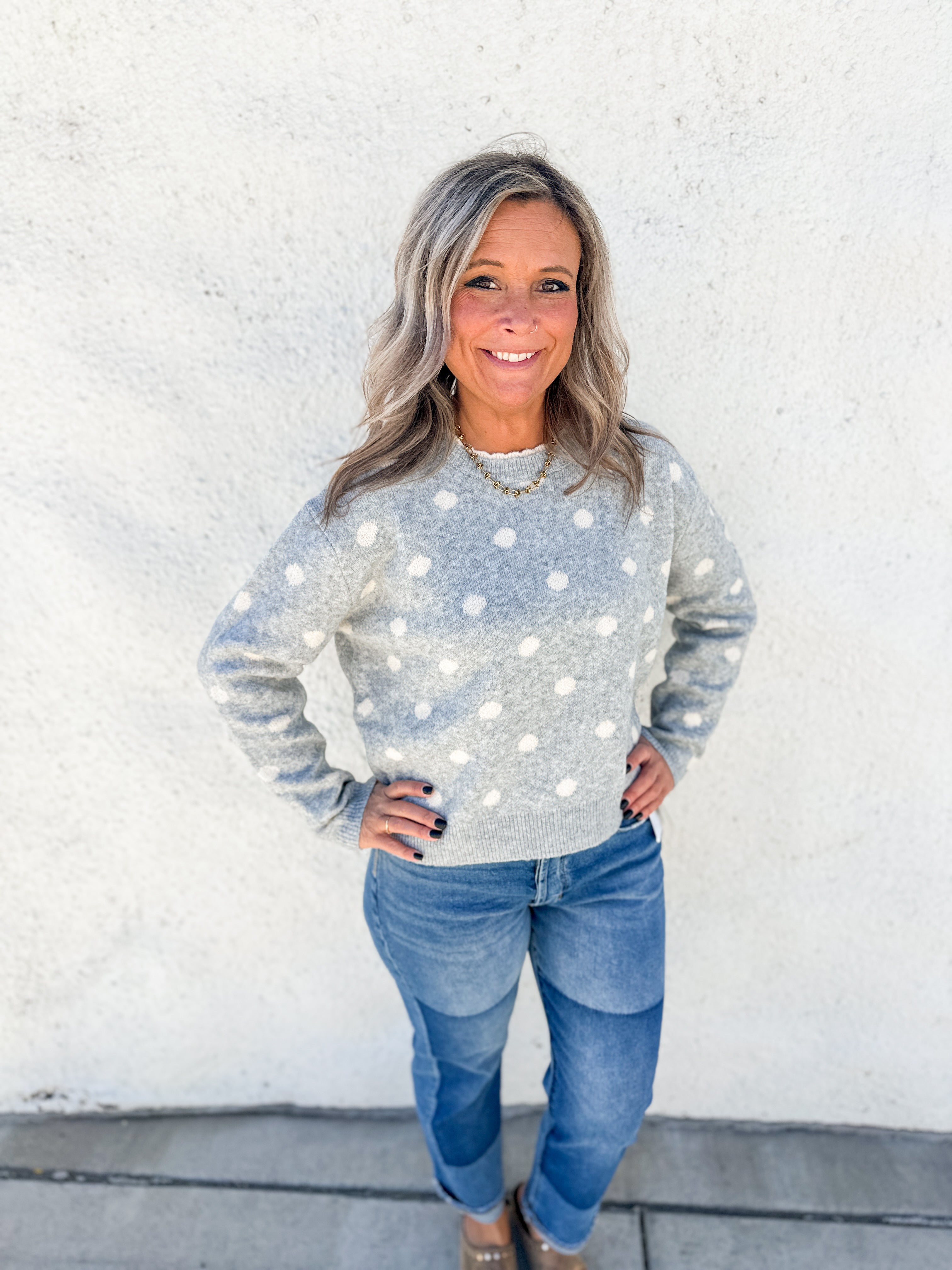 A woman with gray hair stands against a white stucco wall wearing a light gray long-sleeve knit sweater with white polka dots and blue jeans.