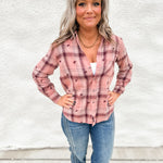 A woman with gray hair stands against a white textured wall wearing a pink and maroon plaid button-up shirt with small embroidered details, paired with blue jeans.
