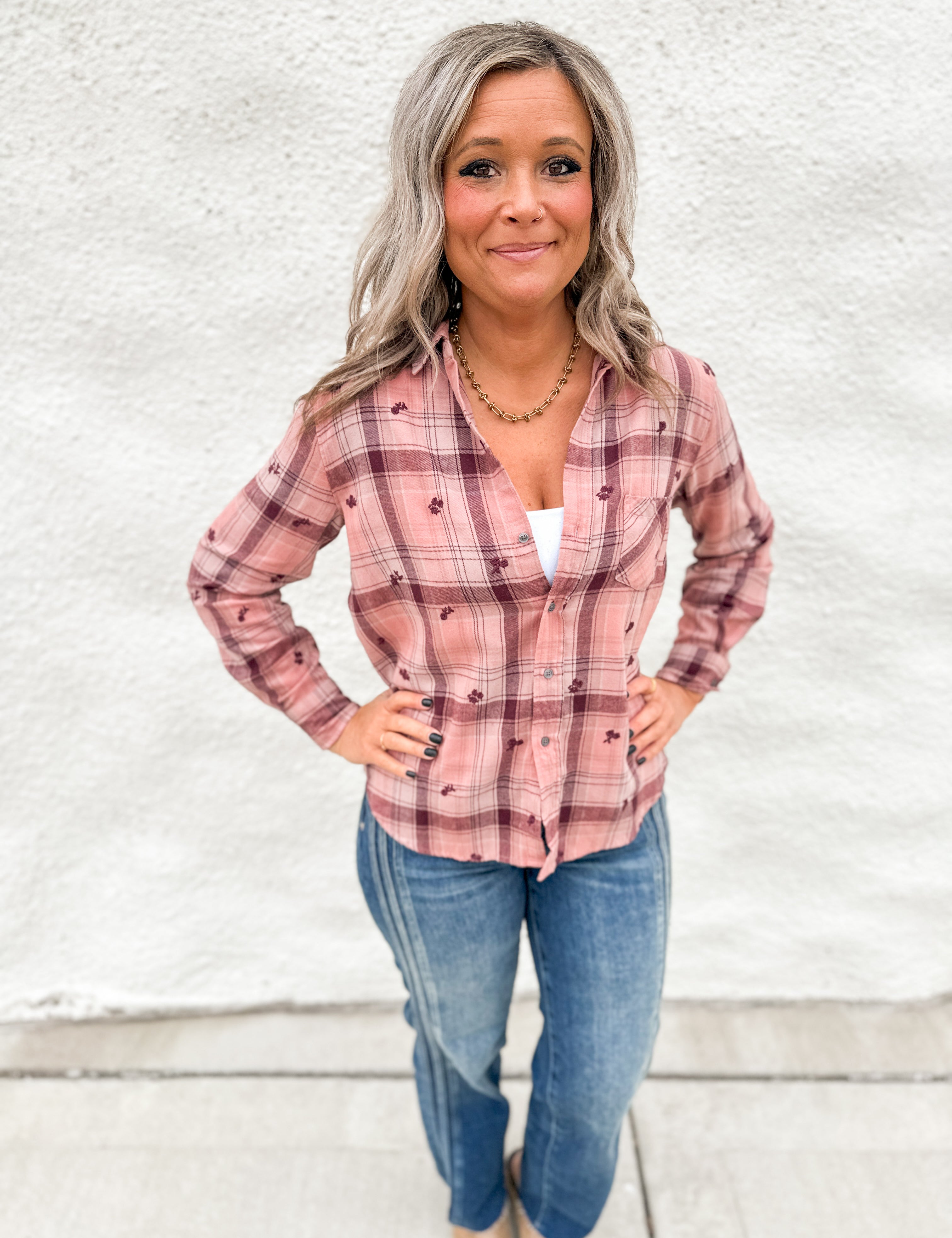 A woman with gray hair stands against a white textured wall wearing a pink and maroon plaid button-up shirt with small embroidered details, paired with blue jeans.