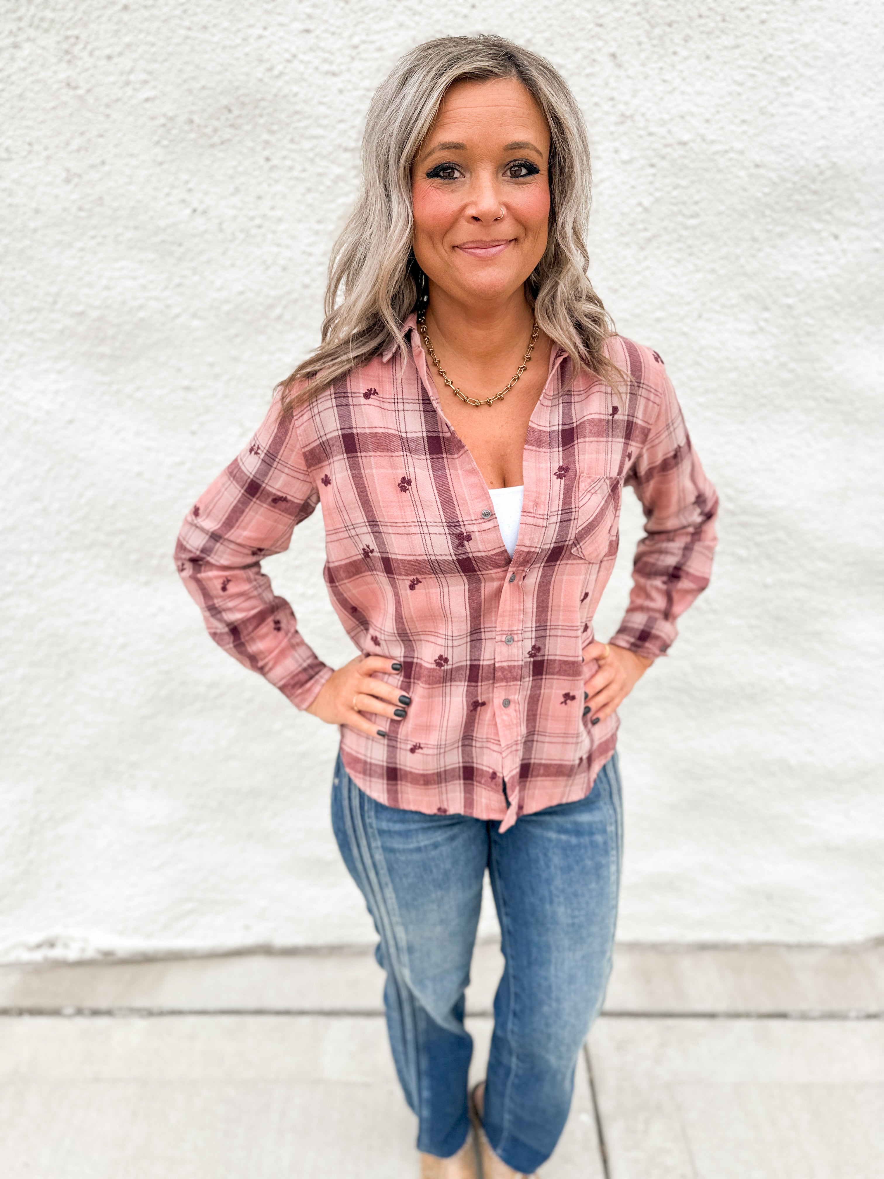 A woman with gray hair stands against a white textured wall wearing a pink and maroon plaid button-up shirt with small embroidered details, paired with blue jeans.