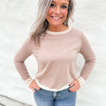 A smiling woman with blonde-highlighted hair wearing a light beige crewneck sweater with cream ribbed trim and blue jeans, standing against a white textured wall.