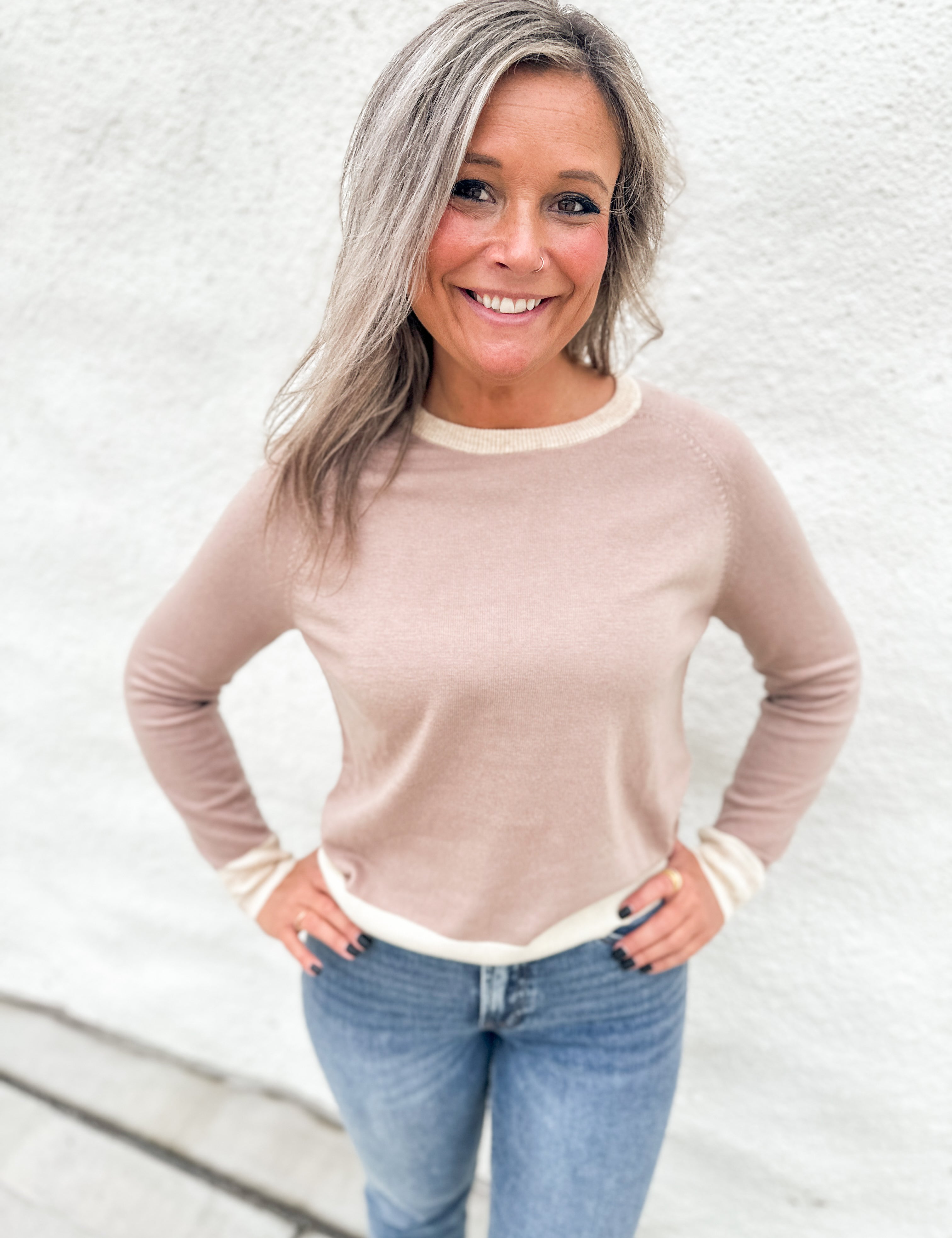 A smiling woman with blonde-highlighted hair wearing a light beige crewneck sweater with cream ribbed trim and blue jeans, standing against a white textured wall.