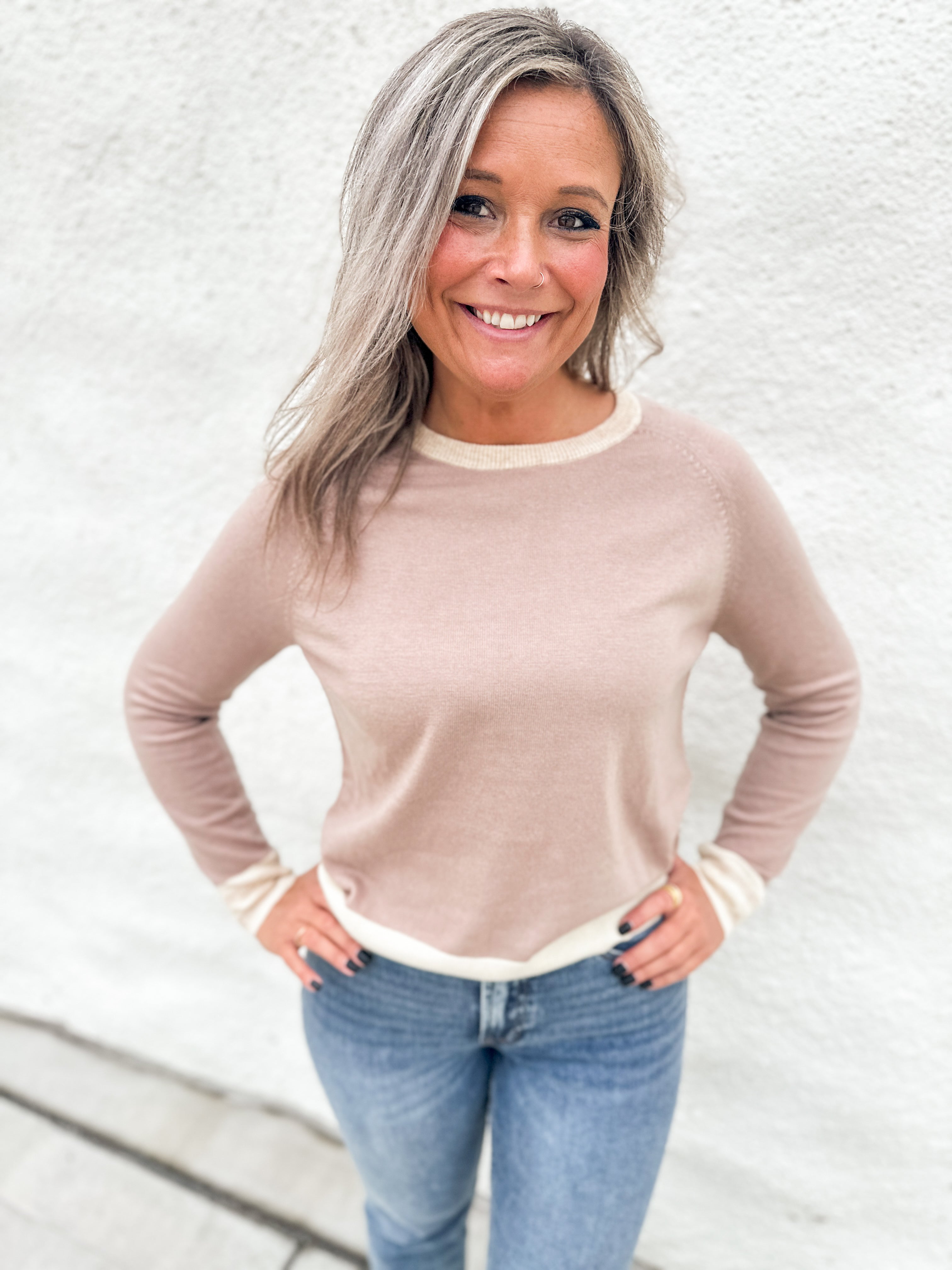 A smiling woman with blonde-highlighted hair wearing a light beige crewneck sweater with cream ribbed trim and blue jeans, standing against a white textured wall.