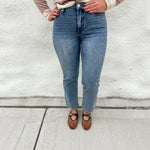 A person wearing straight leg medium-wash mid rise blue jeans by Dear John, a beige long-sleeve sweater, and brown mesh Mary Jane flats, standing against a white stucco wall.