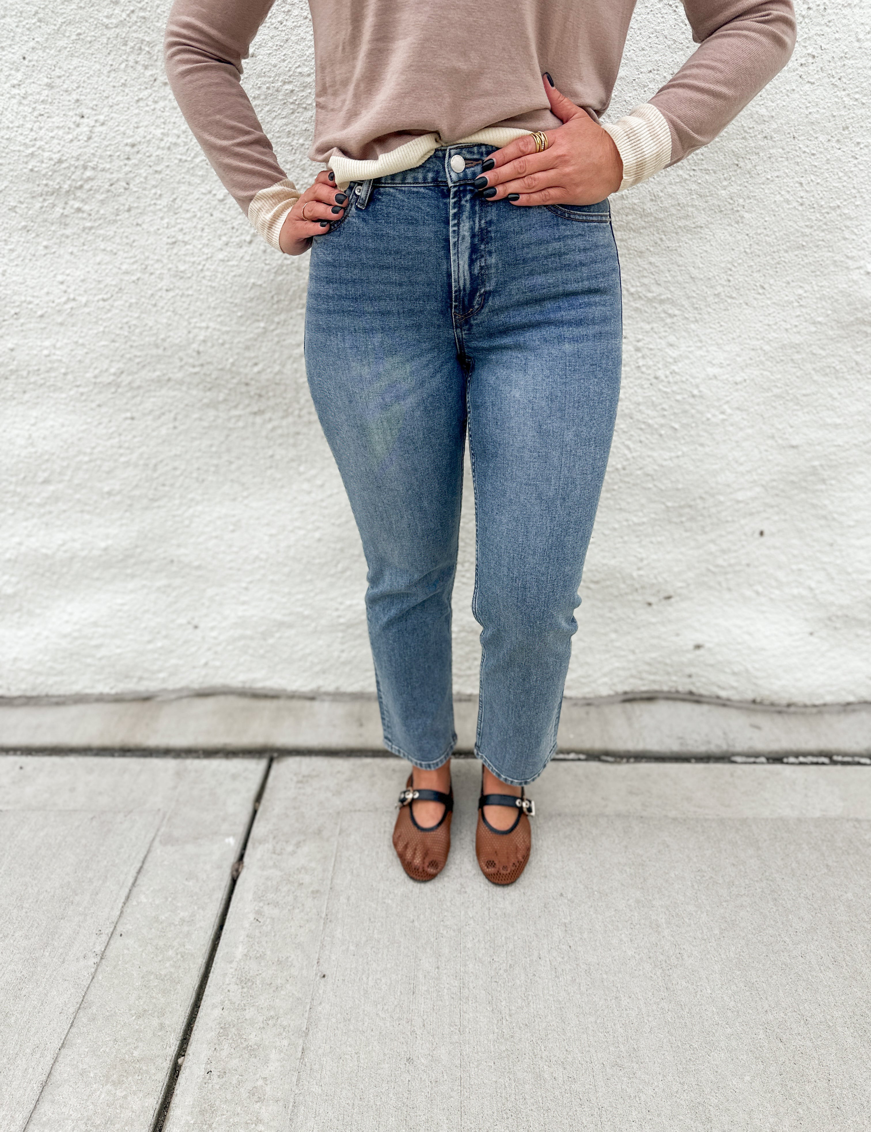 A person wearing straight leg medium-wash mid rise blue jeans by Dear John, a beige long-sleeve sweater, and brown mesh Mary Jane flats, standing against a white stucco wall.