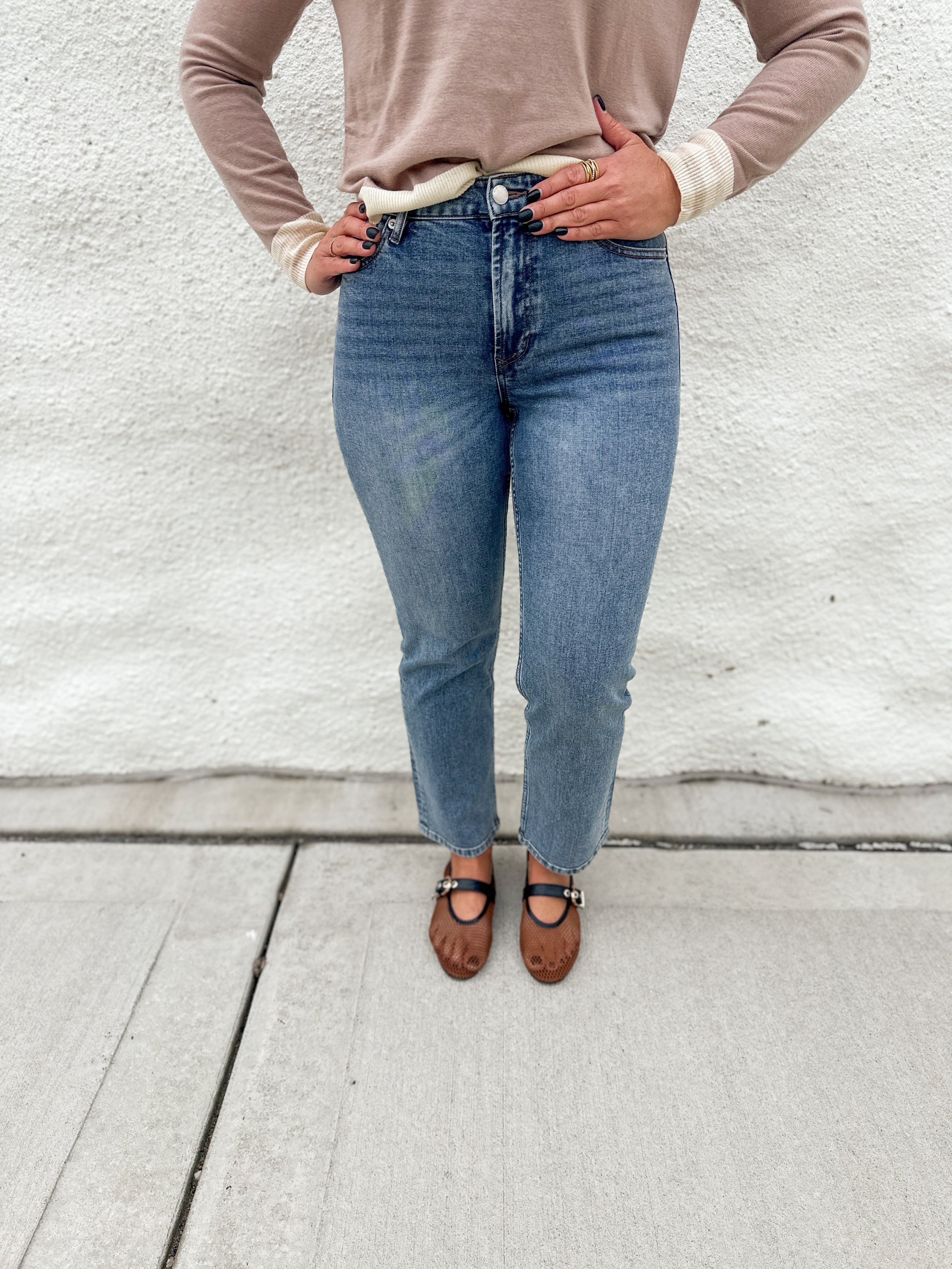 A person wearing straight leg medium-wash mid rise blue jeans by Dear John, a beige long-sleeve sweater, and brown mesh Mary Jane flats, standing against a white stucco wall.