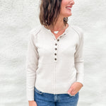 A woman wearing a light beige henley  sweater and blue jeans, standing against a white textured wall.