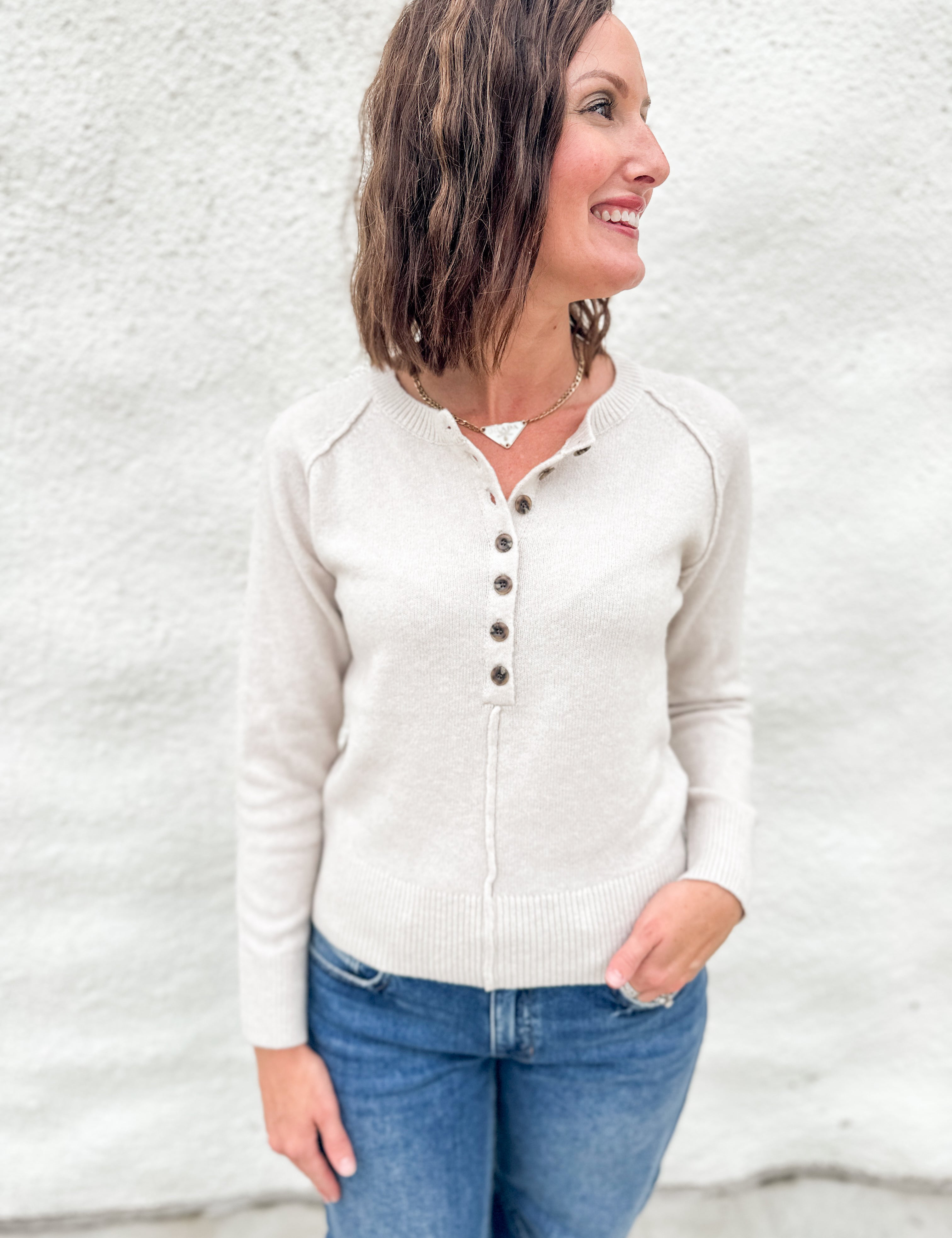 A woman wearing a light beige henley  sweater and blue jeans, standing against a white textured wall.