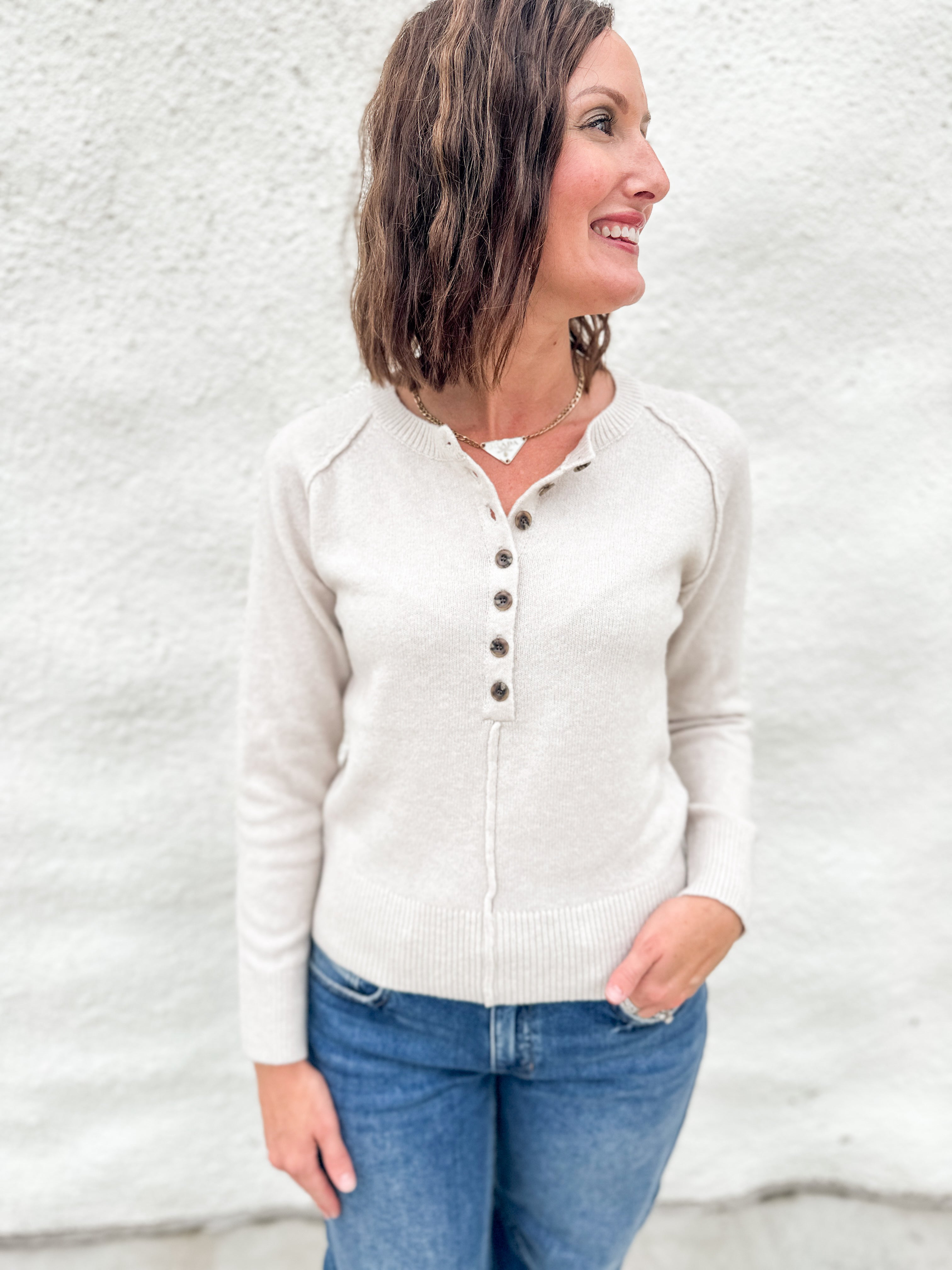 A woman wearing a light beige henley  sweater and blue jeans, standing against a white textured wall.