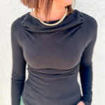 A woman wearing a fitted long-sleeve cowl neck top in black, paired with a light green top or skirt, standing against a light textured wall.