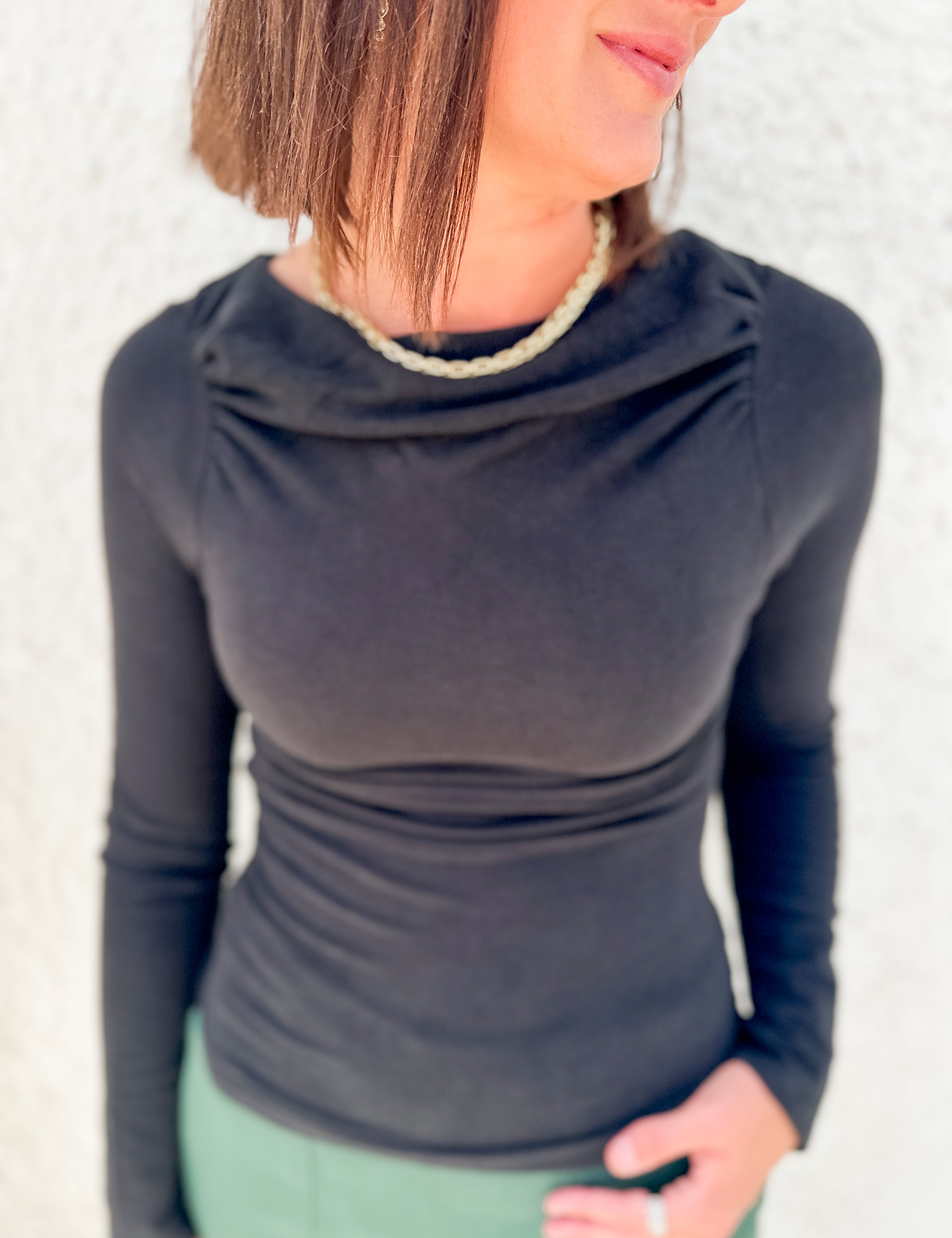 A woman wearing a fitted long-sleeve cowl neck top in black, paired with a light green top or skirt, standing against a light textured wall.