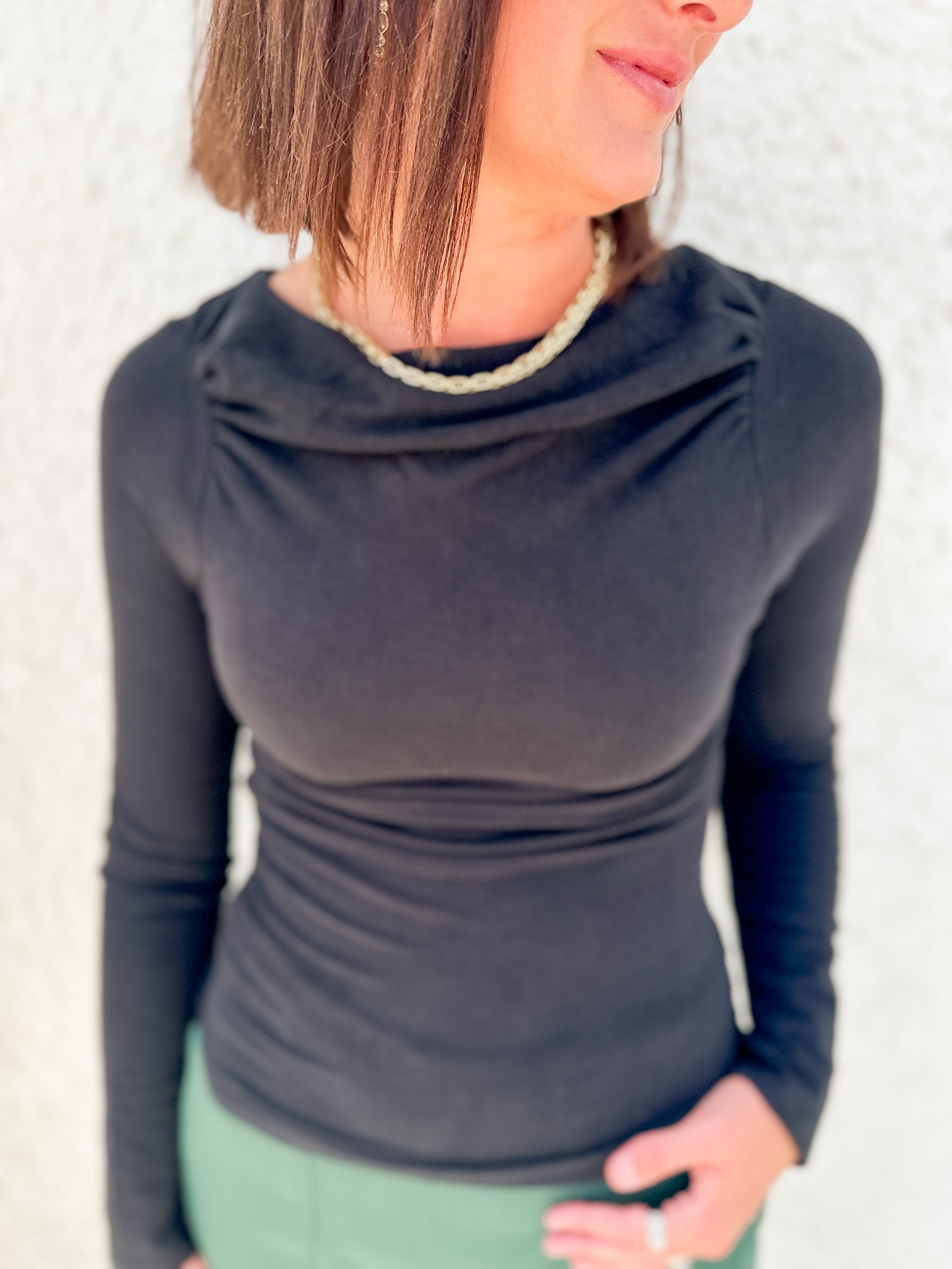 A woman wearing a fitted long-sleeve cowl neck top in black, paired with a light green top or skirt, standing against a light textured wall.