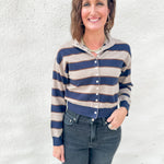 A woman wearing a cropped, long-sleeve button-front cardigan with horizontal navy blue and beige stripes, paired with dark blue jeans, standing against a textured white stucco wall.