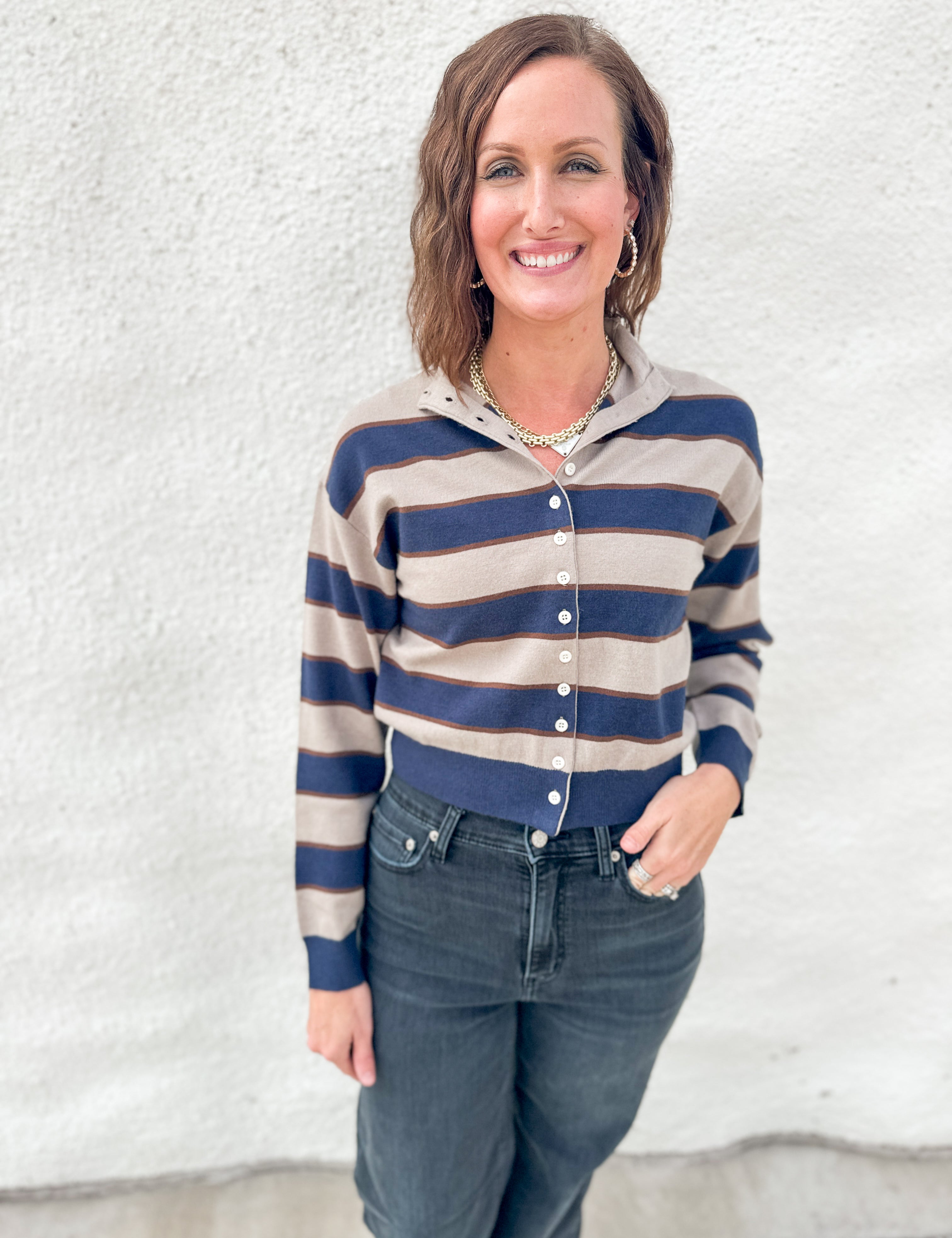 A woman wearing a cropped, long-sleeve button-front cardigan with horizontal navy blue and beige stripes, paired with dark blue jeans, standing against a textured white stucco wall.