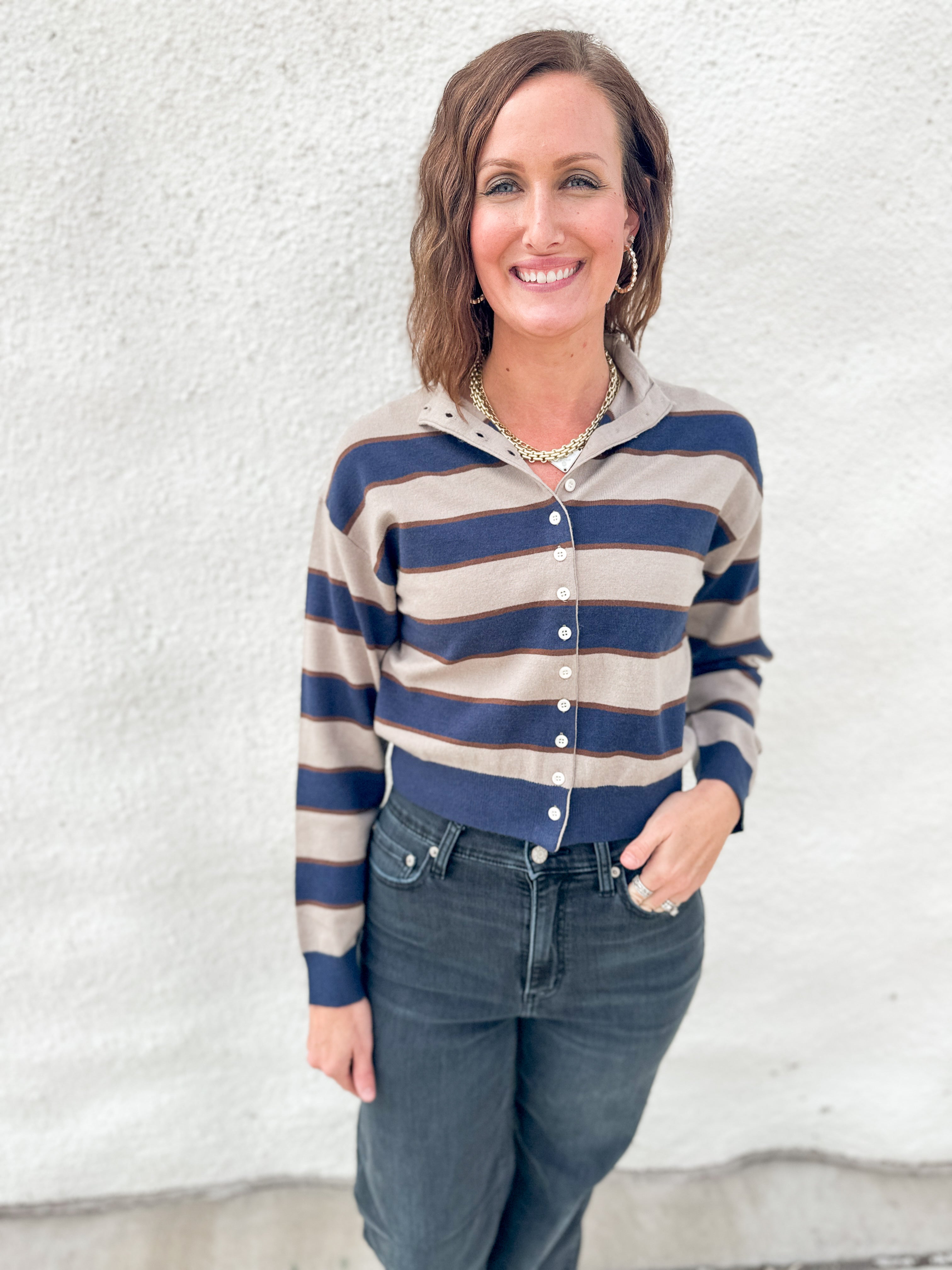 A woman wearing a cropped, long-sleeve button-front cardigan with horizontal navy blue and beige stripes, paired with dark blue jeans, standing against a textured white stucco wall.