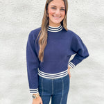 A woman modeling a navy blue mock neck sweater with white striped ribbed trim, paired with blue jeans, standing against a textured white wall.