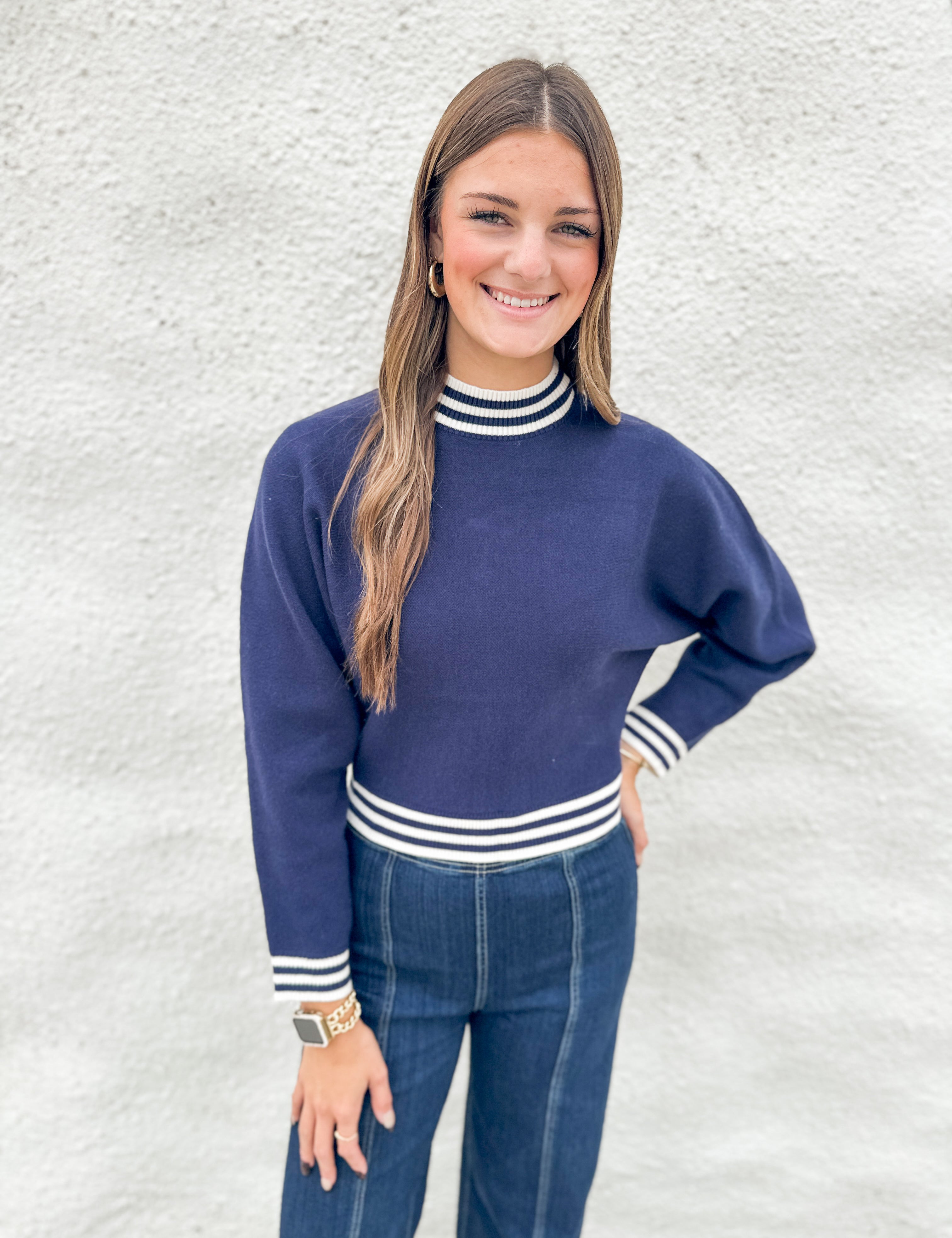 A woman modeling a navy blue mock neck sweater with white striped ribbed trim, paired with blue jeans, standing against a textured white wall.