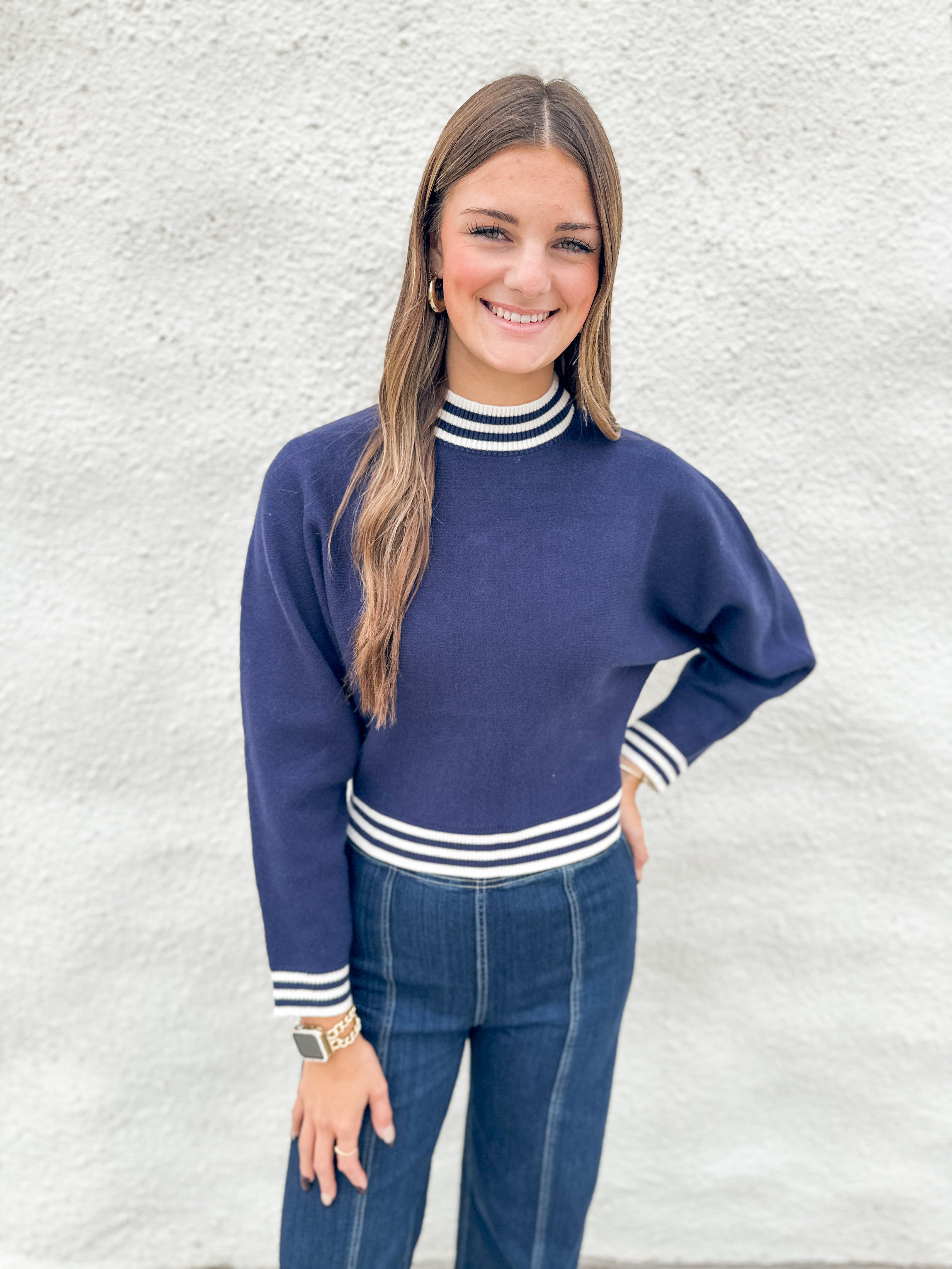 A woman modeling a navy blue mock neck sweater with white striped ribbed trim, paired with blue jeans, standing against a textured white wall.