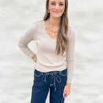 A woman modeling a beige cable-knit long-sleeve sweater with a scalloped V-neckline, paired with dark blue jeans and gold jewelry, standing against a white textured wall.