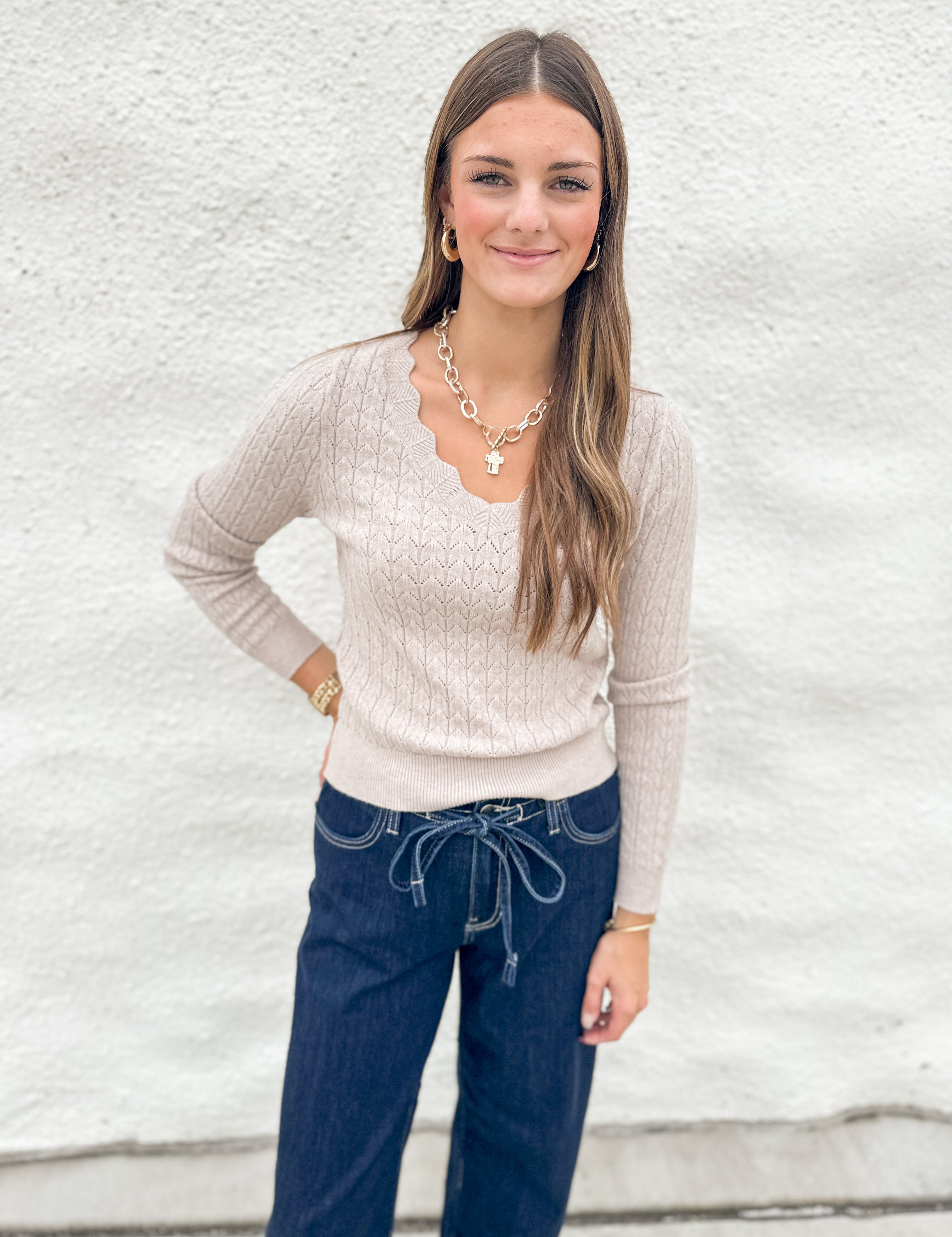 A woman modeling a beige cable-knit long-sleeve sweater with a scalloped V-neckline, paired with dark blue jeans and gold jewelry, standing against a white textured wall.