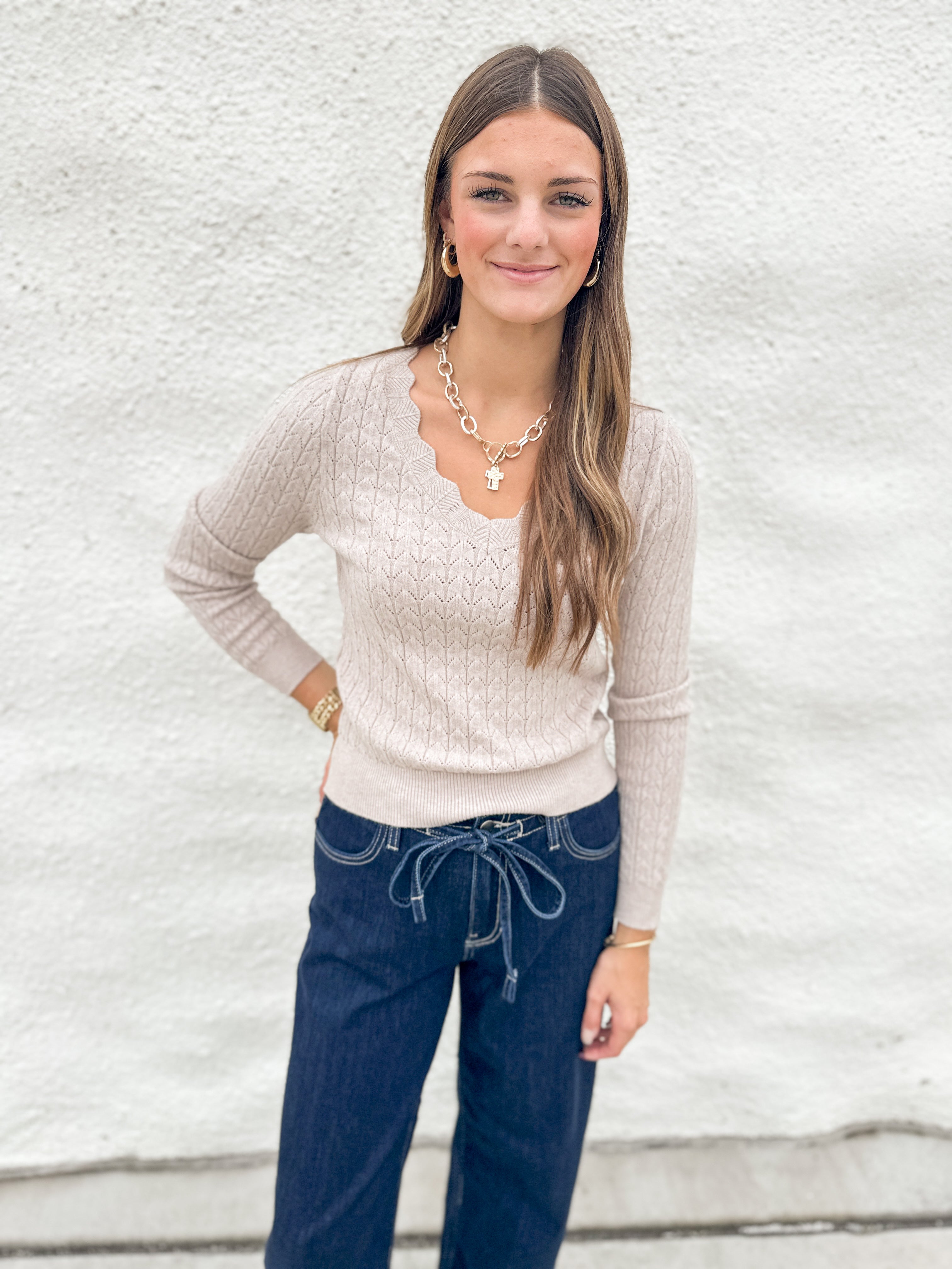 A woman modeling a beige cable-knit long-sleeve sweater with a scalloped V-neckline, paired with dark blue jeans and gold jewelry, standing against a white textured wall.