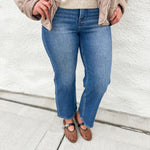 A person wearing blue wide-leg jeans, a taupe quilted jacket, a dark gray top, and brown mesh Mary Jane flats, standing against a white stucco wall.