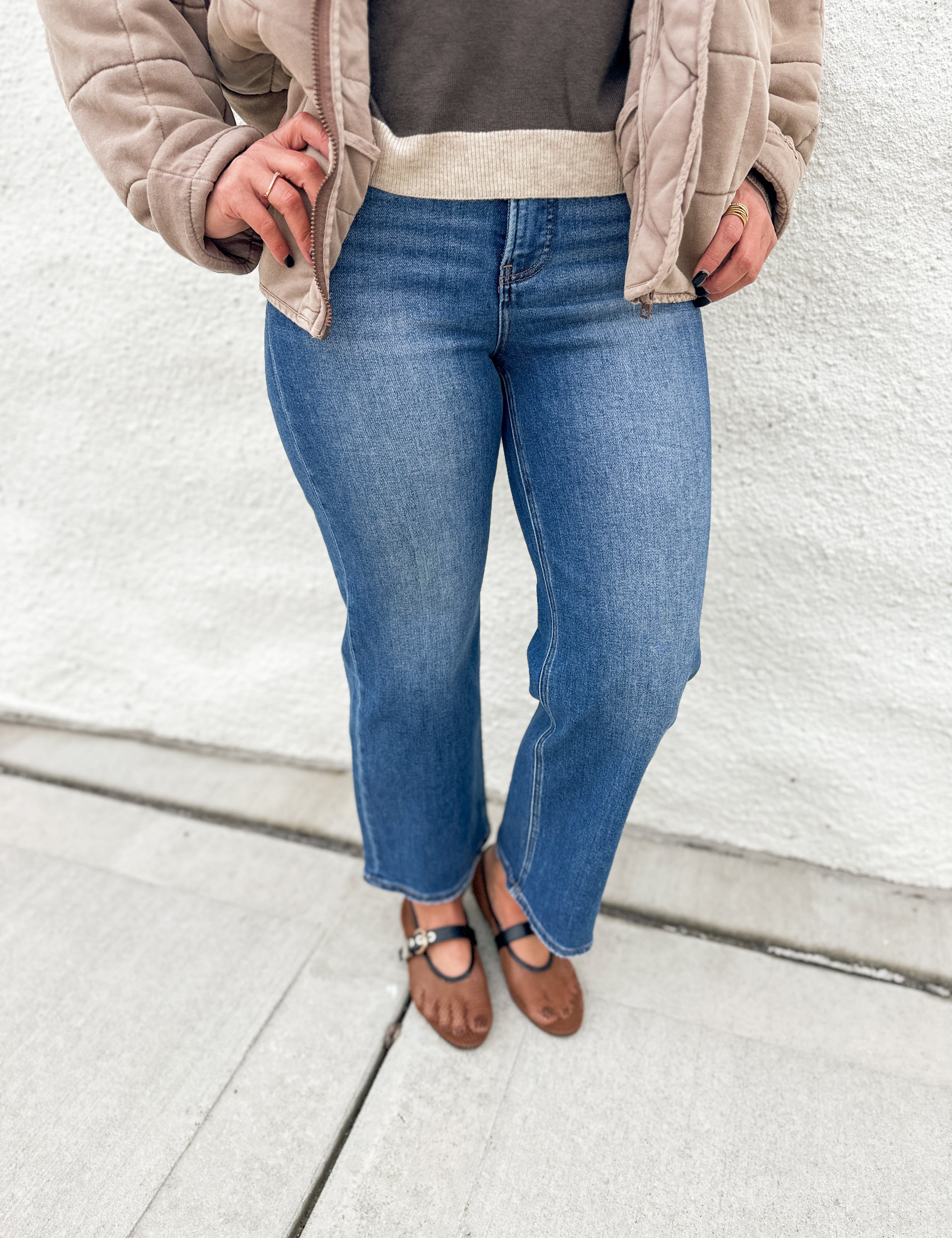 A person wearing blue wide-leg jeans, a taupe quilted jacket, a dark gray top, and brown mesh Mary Jane flats, standing against a white stucco wall.