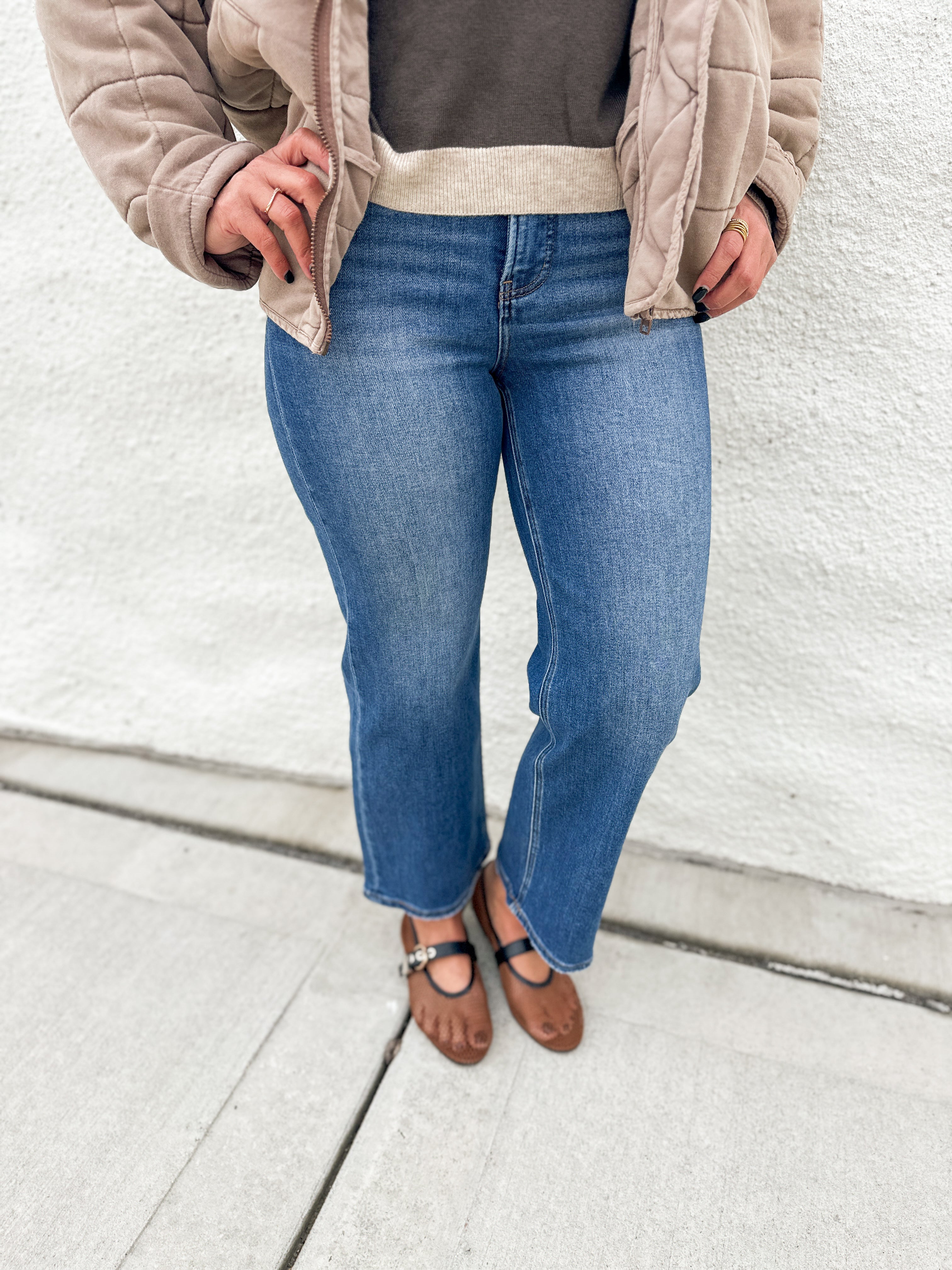 A person wearing blue wide-leg jeans, a taupe quilted jacket, a dark gray top, and brown mesh Mary Jane flats, standing against a white stucco wall.