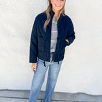A woman stands against a white stucco wall wearing a black quilted bomber jacket over a gray and white striped shirt, blue jeans, and beige pointed-toe flats.