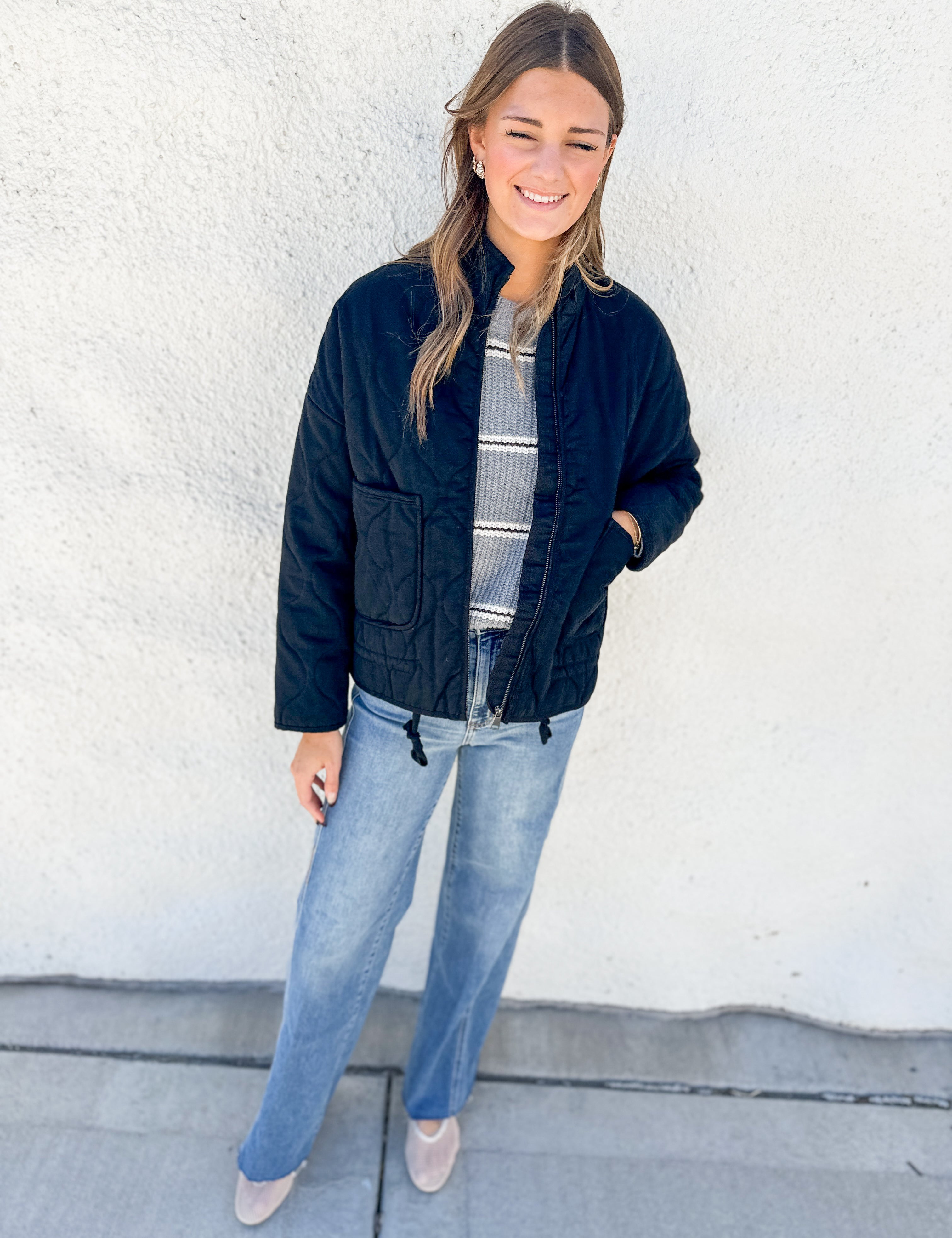 A woman stands against a white stucco wall wearing a black quilted bomber jacket over a gray and white striped shirt, blue jeans, and beige pointed-toe flats.