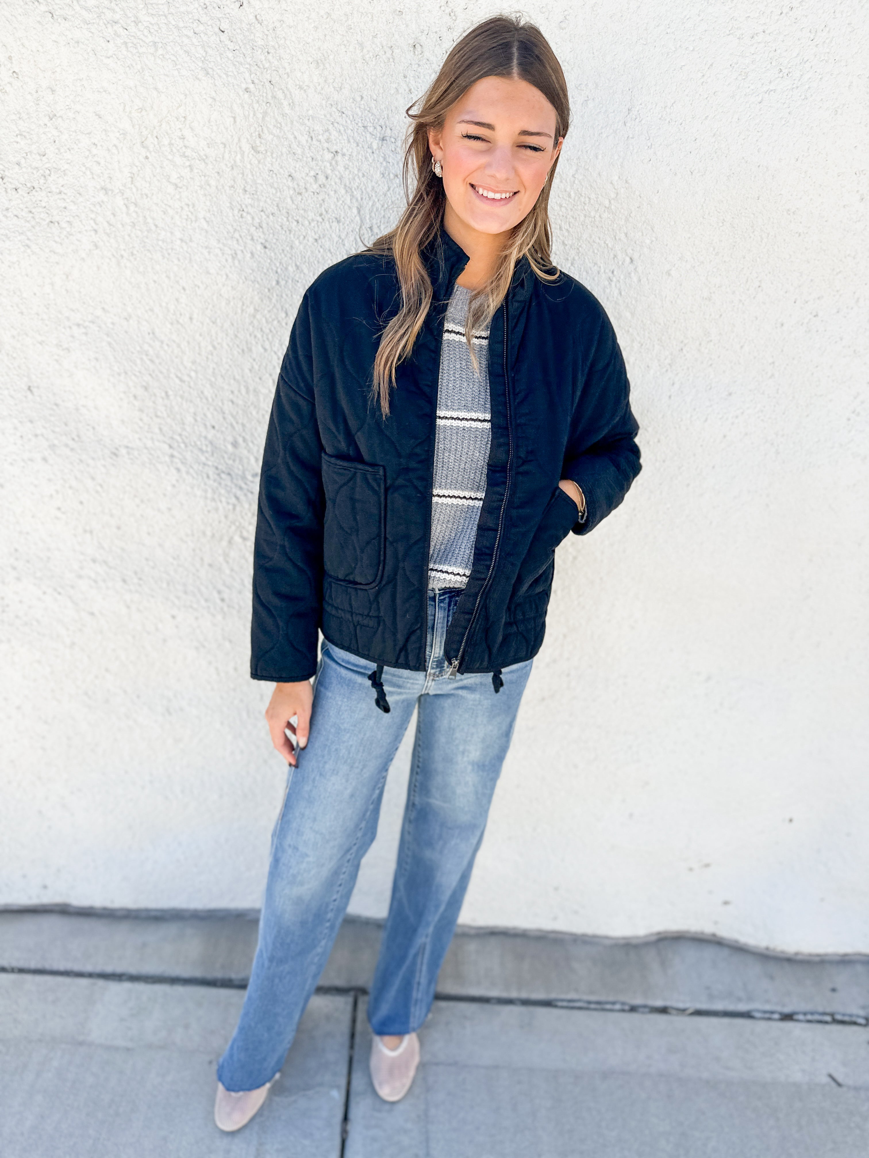 A woman stands against a white stucco wall wearing a black quilted bomber jacket over a gray and white striped shirt, blue jeans, and beige pointed-toe flats.