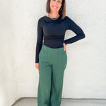 A woman modeling a black long-sleeve cowl-neck top and green wide-leg pants, standing against a textured white stucco wall.