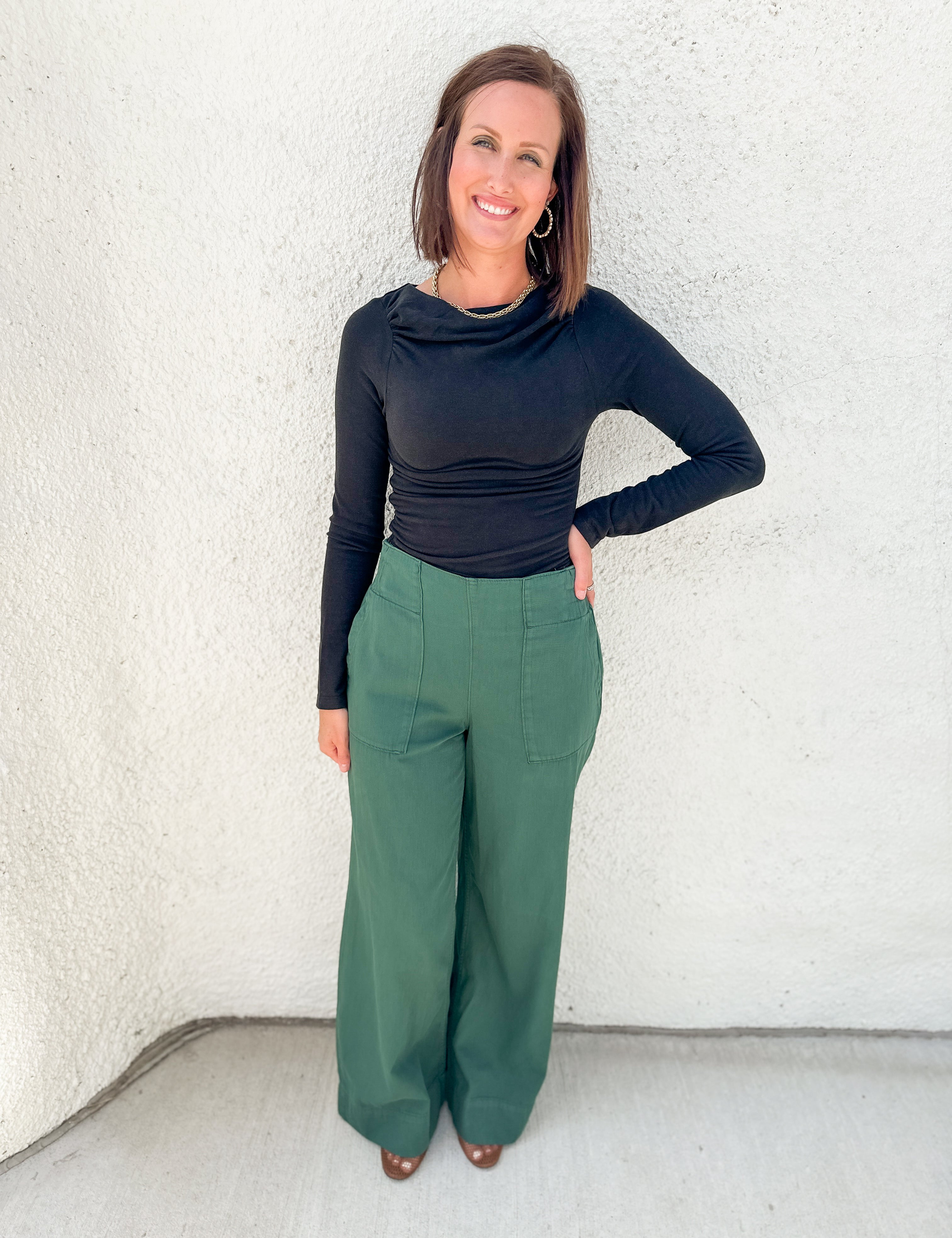 A woman modeling a black long-sleeve cowl-neck top and green wide-leg pants, standing against a textured white stucco wall.