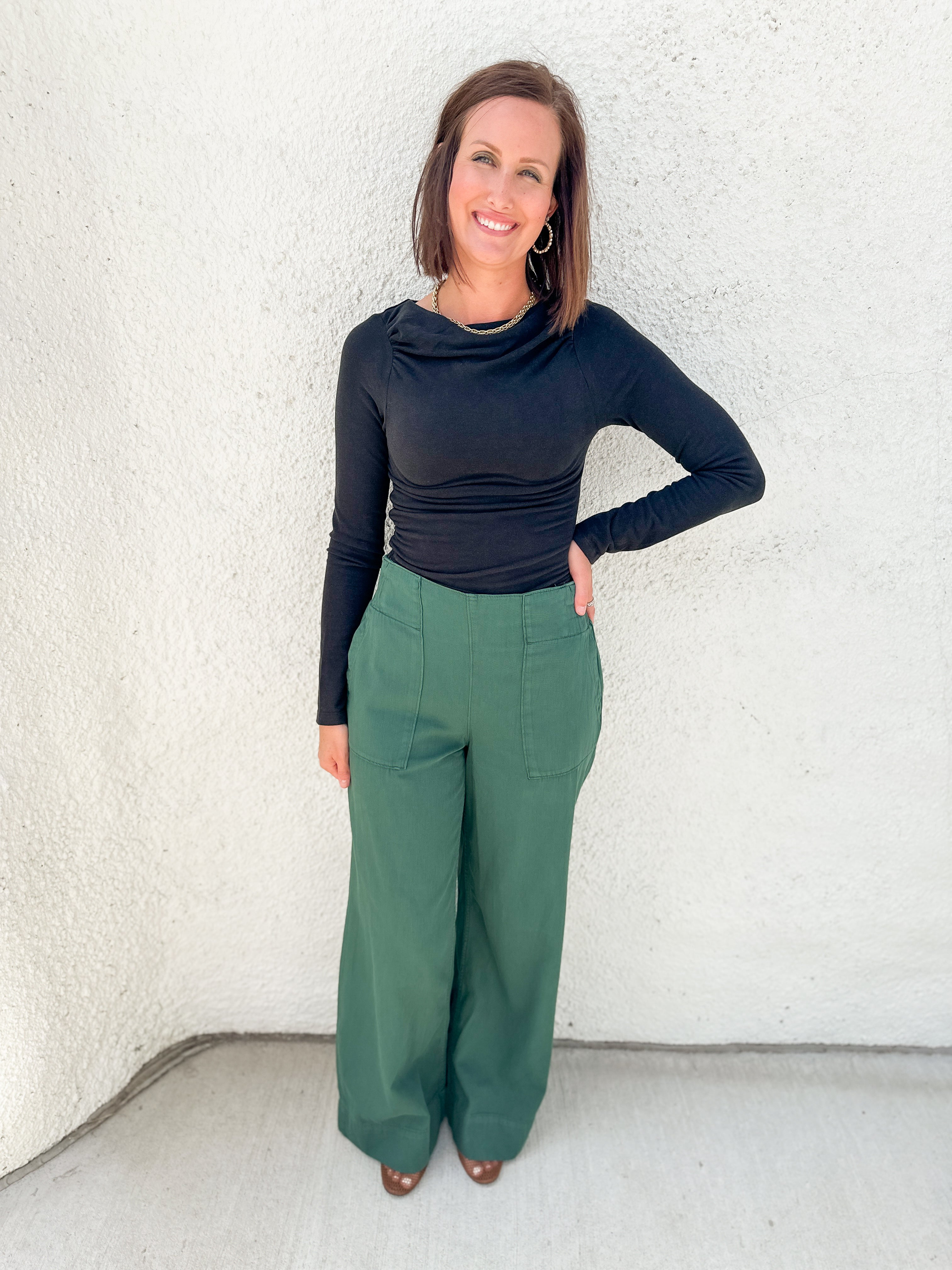 A woman modeling a black long-sleeve cowl-neck top and green wide-leg pants, standing against a textured white stucco wall.