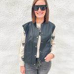 A woman wearing a black faux-leather bomber jacket with gold buttons over a cream knit sweater, paired with gray jeans and black sunglasses, standing against a white textured wall.