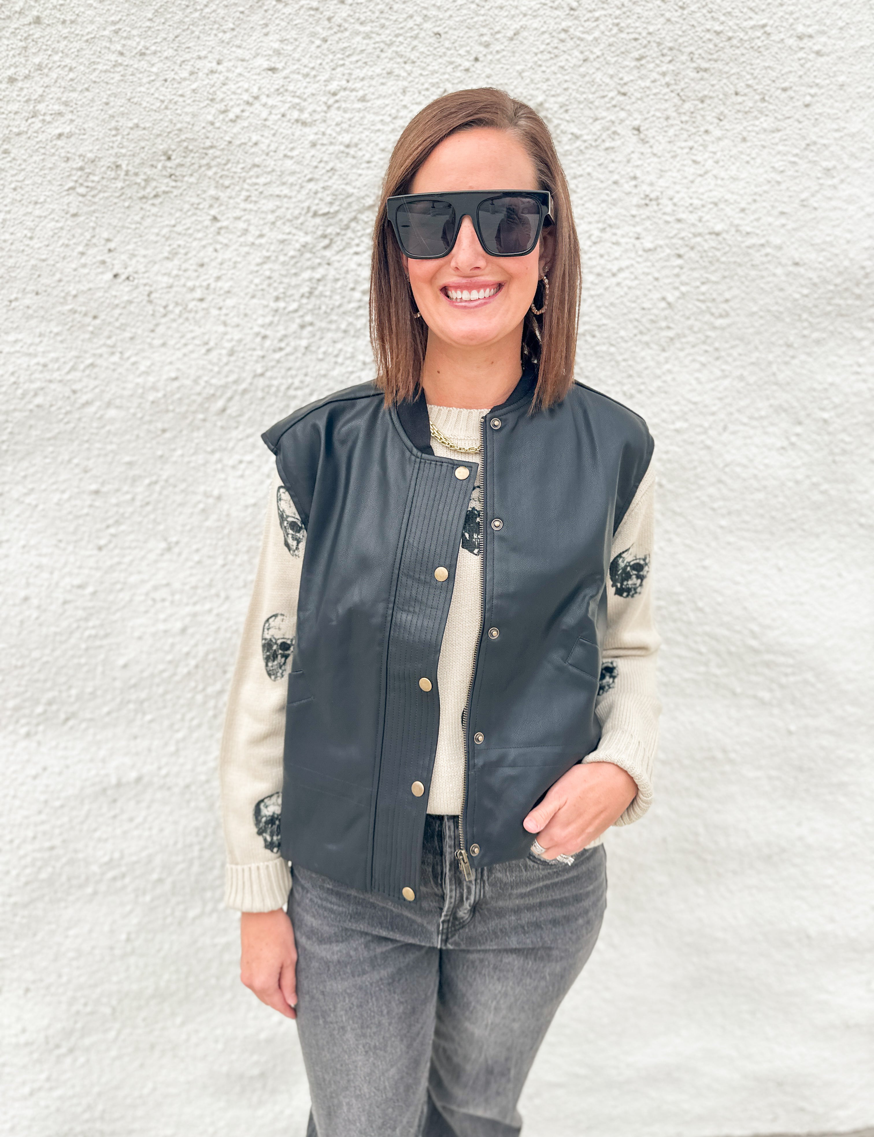 A woman wearing a black faux-leather bomber jacket with gold buttons over a cream knit sweater, paired with gray jeans and black sunglasses, standing against a white textured wall.