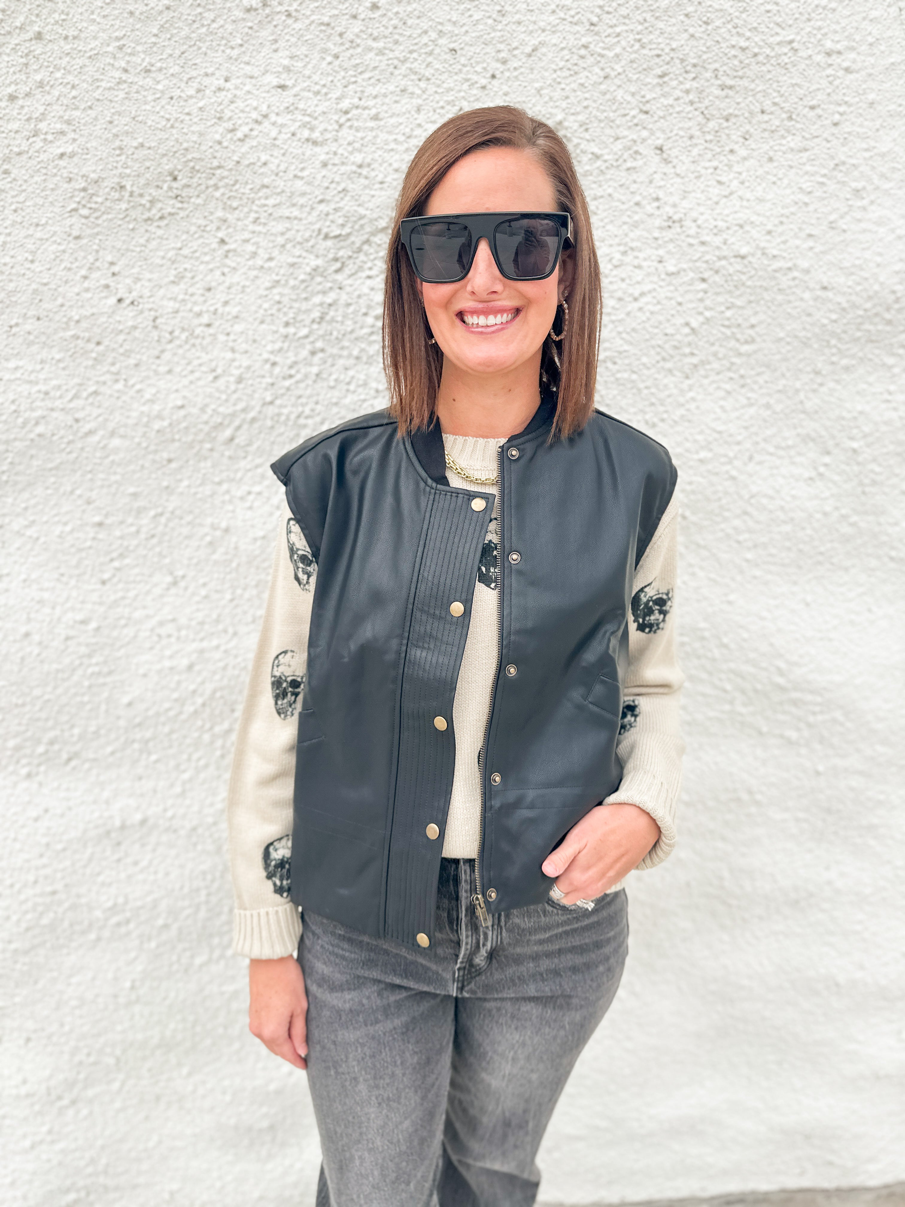 A woman wearing a black faux-leather bomber jacket with gold buttons over a cream knit sweater, paired with gray jeans and black sunglasses, standing against a white textured wall.