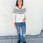 A woman stands outdoors against a white stucco wall wearing a cream knit half-zip sweater with a dark gray Nordic/Fair Isle yoke pattern, blue jeans, and black shoes.