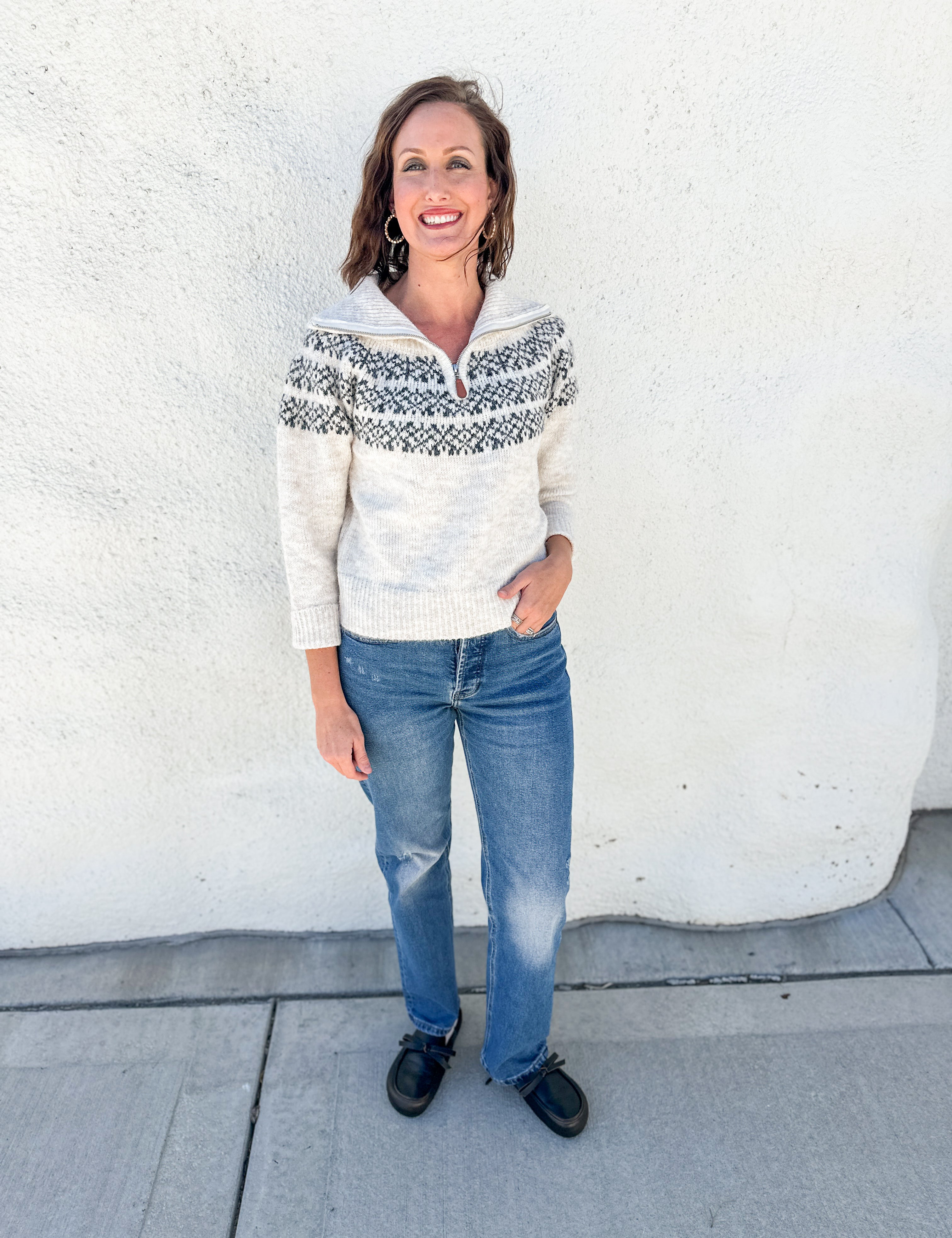 A woman stands outdoors against a white stucco wall wearing a cream knit half-zip sweater with a dark gray Nordic/Fair Isle yoke pattern, blue jeans, and black shoes.