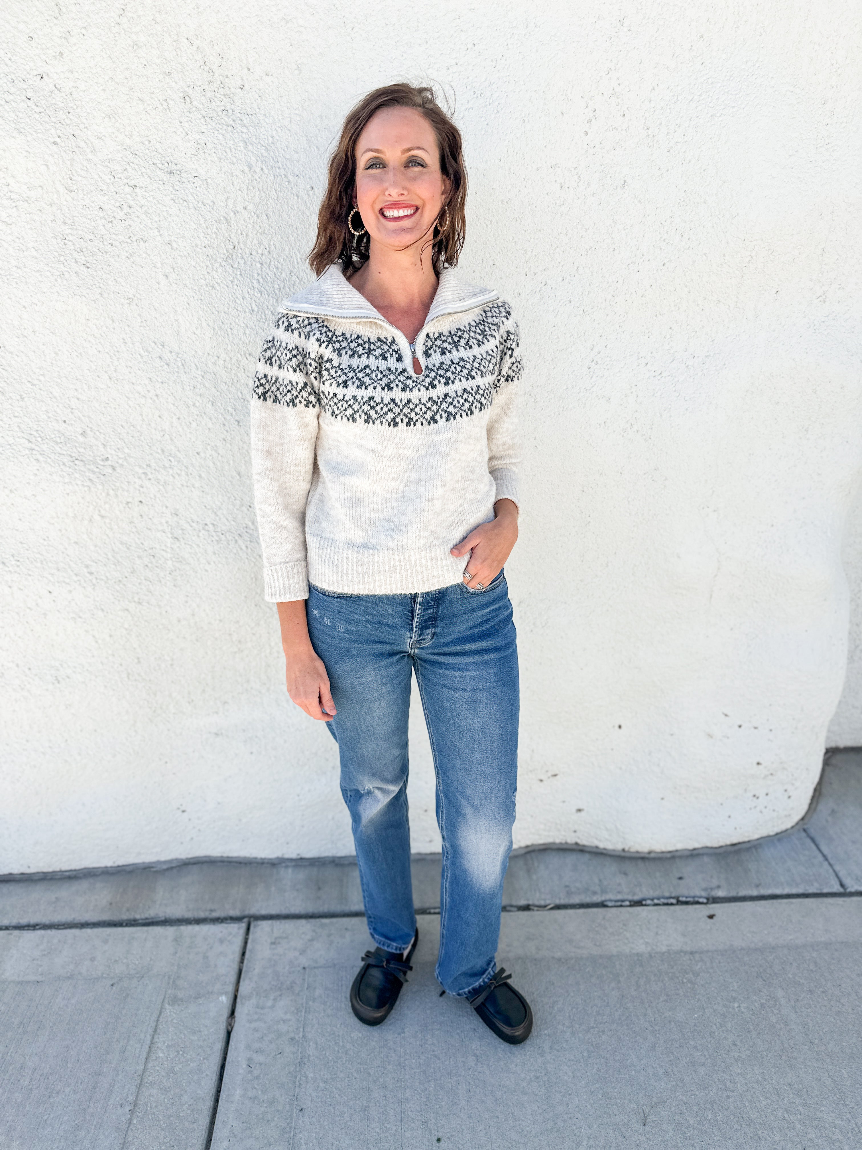 A woman stands outdoors against a white stucco wall wearing a cream knit half-zip sweater with a dark gray Nordic/Fair Isle yoke pattern, blue jeans, and black shoes.