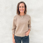 A woman smiling and posing in front of a white textured wall, wearing a beige ribbed knit long-sleeve sweater and dark gray jeans.