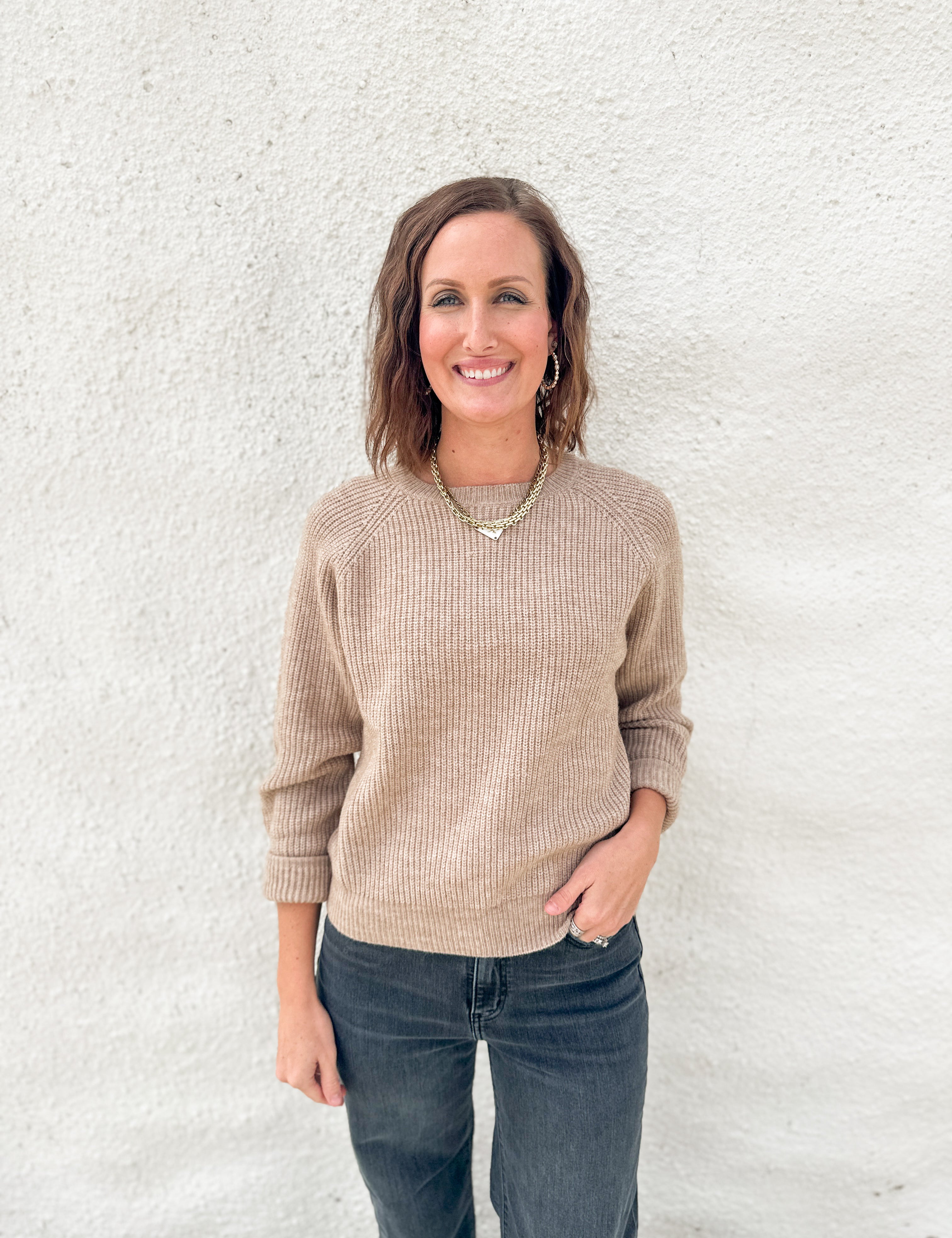 A woman smiling and posing in front of a white textured wall, wearing a beige ribbed knit long-sleeve sweater and dark gray jeans.