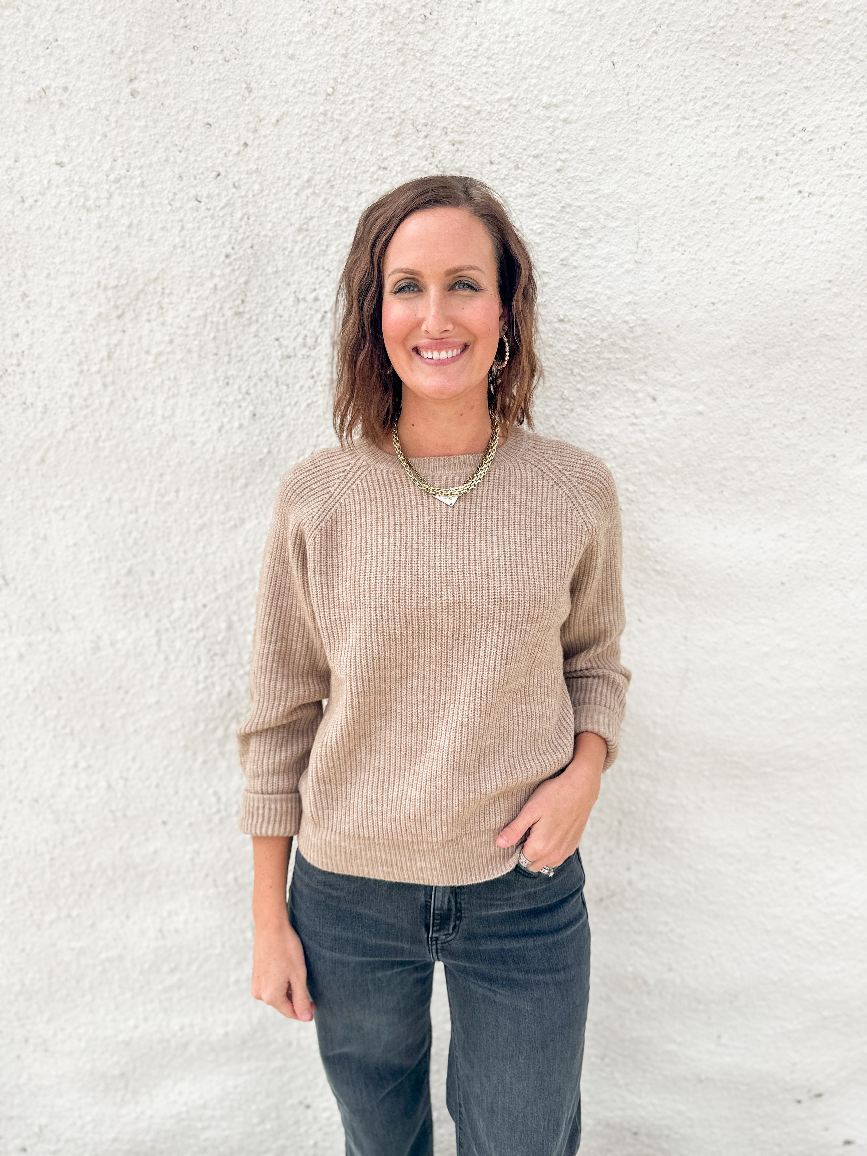 A woman smiling and posing in front of a white textured wall, wearing a beige ribbed knit long-sleeve sweater and dark gray jeans.