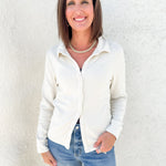 A woman with shoulder-length brown hair smiling, wearing a cream-colored long-sleeve button-down shirt and blue jeans, standing against a textured white stucco wall.