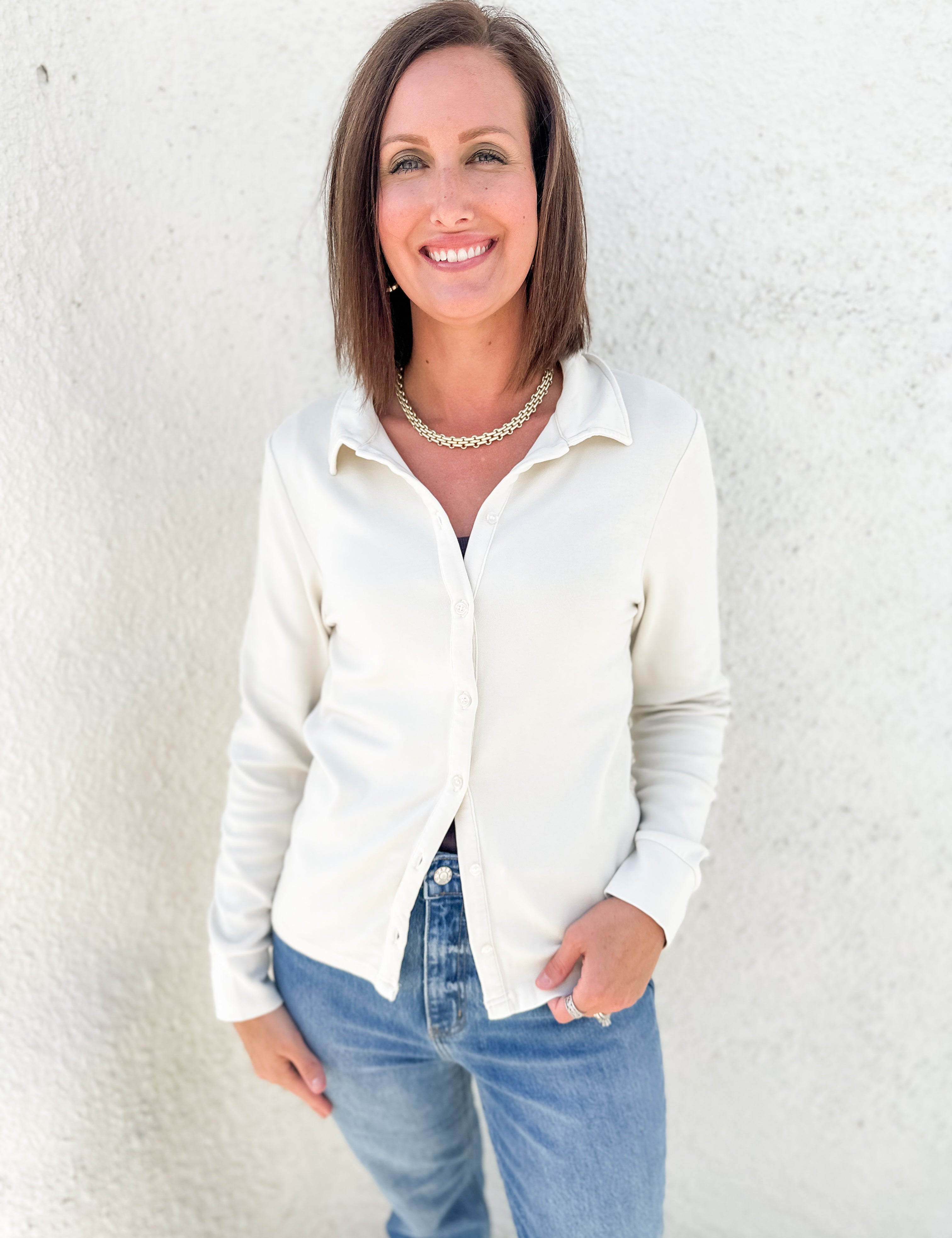 A woman with shoulder-length brown hair smiling, wearing a cream-colored long-sleeve button-down shirt and blue jeans, standing against a textured white stucco wall.