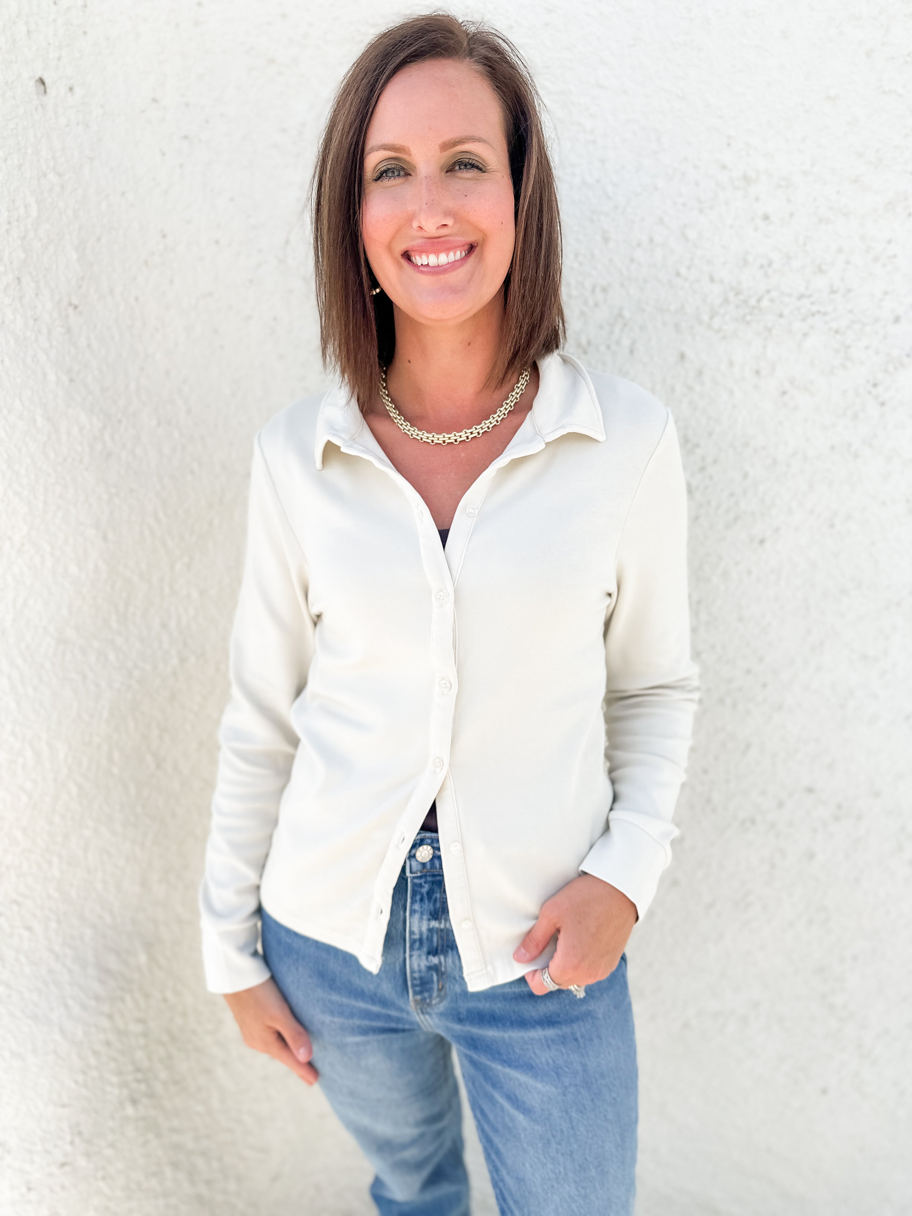 A woman with shoulder-length brown hair smiling, wearing a cream-colored long-sleeve button-down shirt and blue jeans, standing against a textured white stucco wall.