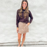 A woman stands against a white stucco wall wearing a dark brown long-sleeve knit sweater, a beige fawn print denim mini skirt, and cream mesh mary jane flats.