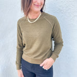 A woman wearing an olive green long-sleeve crewneck sweater and dark navy pants, standing against a light gray textured wall.