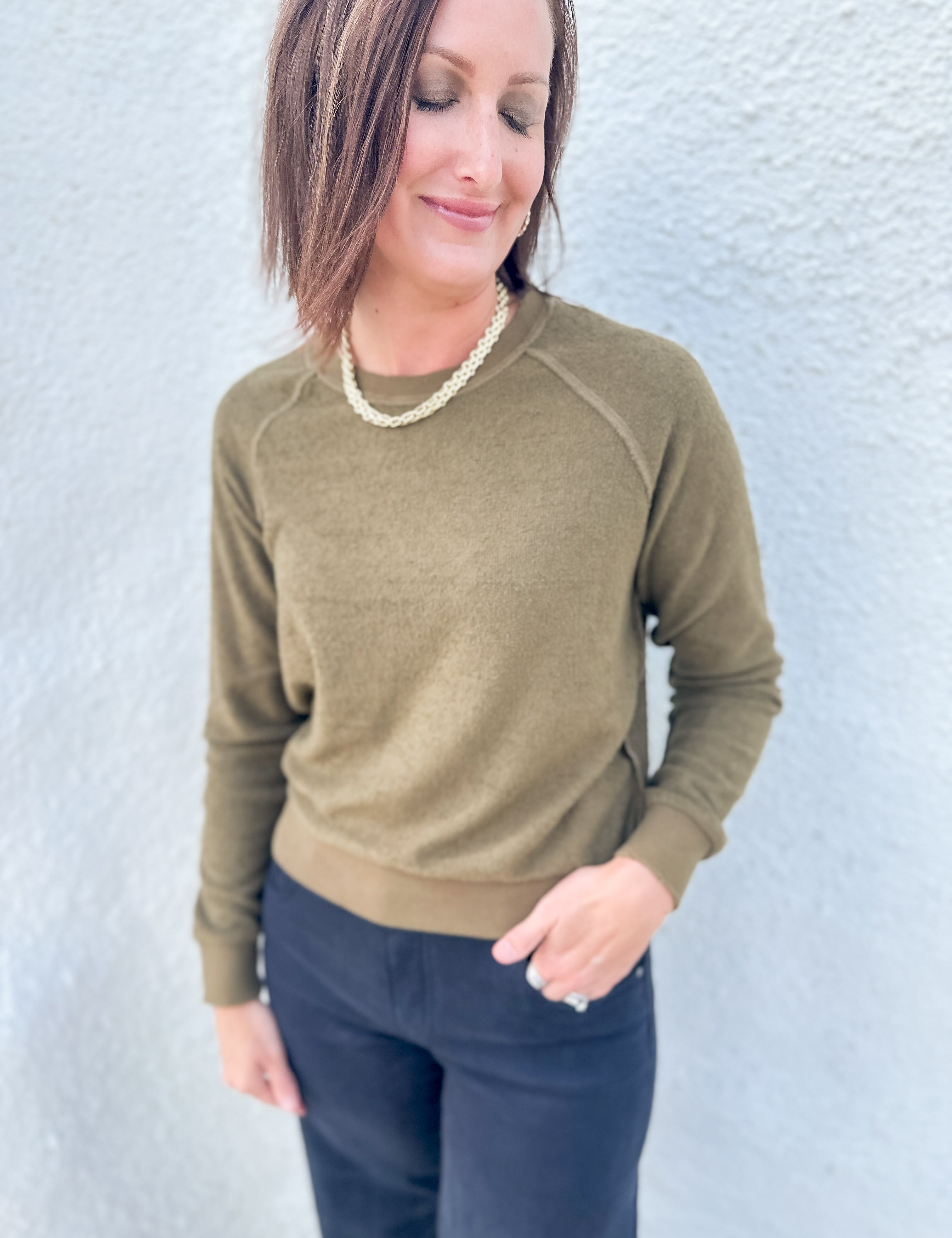 A woman wearing an olive green long-sleeve crewneck sweater and dark navy pants, standing against a light gray textured wall.